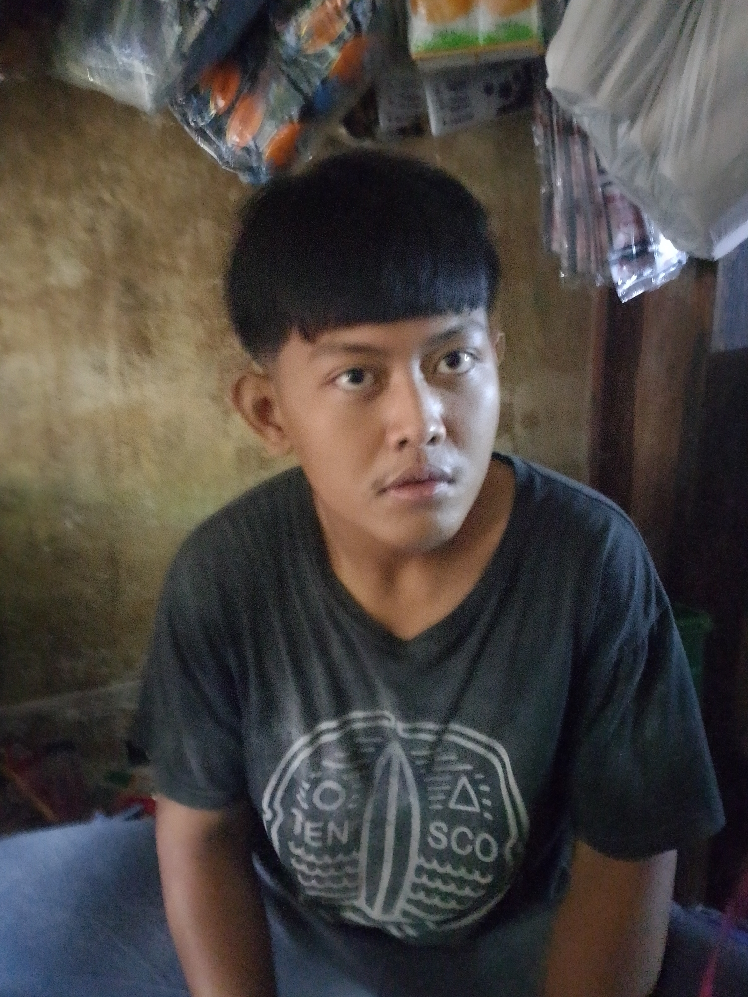 A young male with short, dark hair is pictured in a close-up shot. He is wearing a dark grey t-shirt with a white graphic design on the front. The graphic appears to be circular and contains symbols and text that might be "TEN" and "SCO". His gaze is directed towards the camera. The background is blurred and consists of a textured wall on the left and what appears to be shelves with various items, possibly in a shop, on the right. The lighting suggests an indoor environment. Based on the context provided, this image was taken in Palembang, Indonesia.