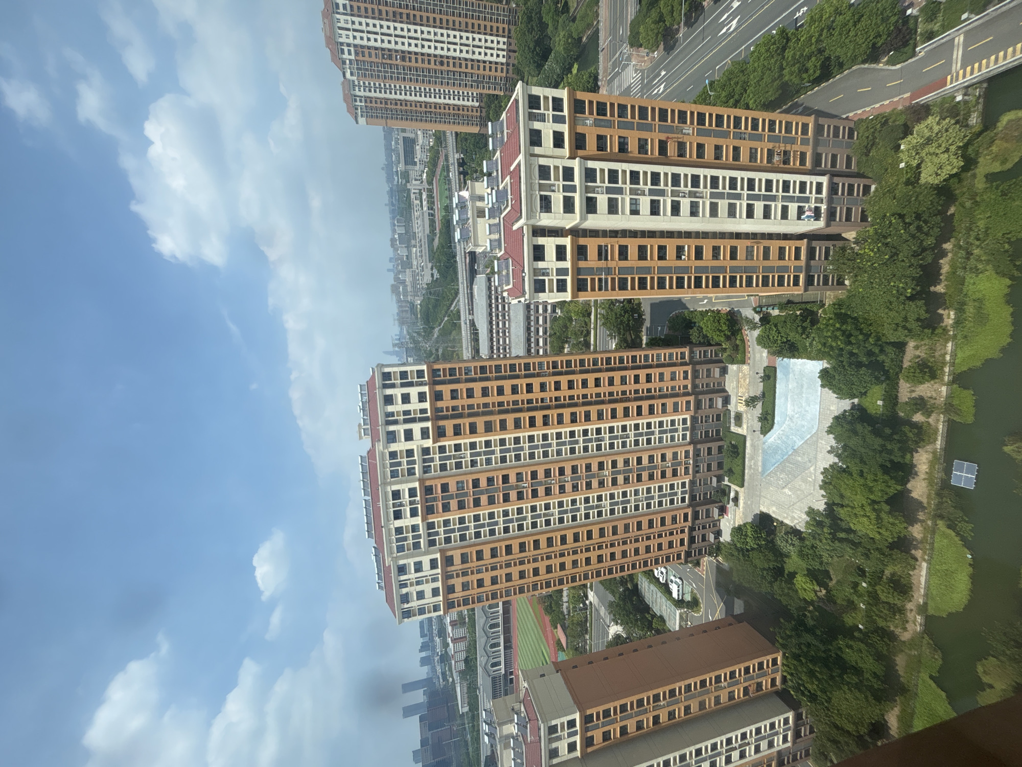 An aerial, high-angle perspective showcases a modern urban landscape in Suzhou Industrial Park, China, bathed in bright daylight under a partly cloudy sky. The scene is dominated by several tall, multi-story residential buildings with a distinctive architectural style, featuring a combination of orange and off-white facades punctuated by rows of windows. These structures appear to be part of a planned community, surrounded by lush green trees and manicured landscaping.
In the foreground, a tranquil body of water, possibly a pond or canal, reflects the sky and surrounding greenery. A single, floating solar panel is visible on the water's surface. Beyond the immediate residential blocks, the view extends to a sports field with a track, and further into the distance, a sprawling cityscape with numerous other buildings and infrastructure, including roads, bridges, and what appears to be industrial or commercial complexes. The overall impression is one of a well-developed and modern urban environment with a blend of residential, recreational, and commercial elements. There are no visible individuals or animals in the frame.