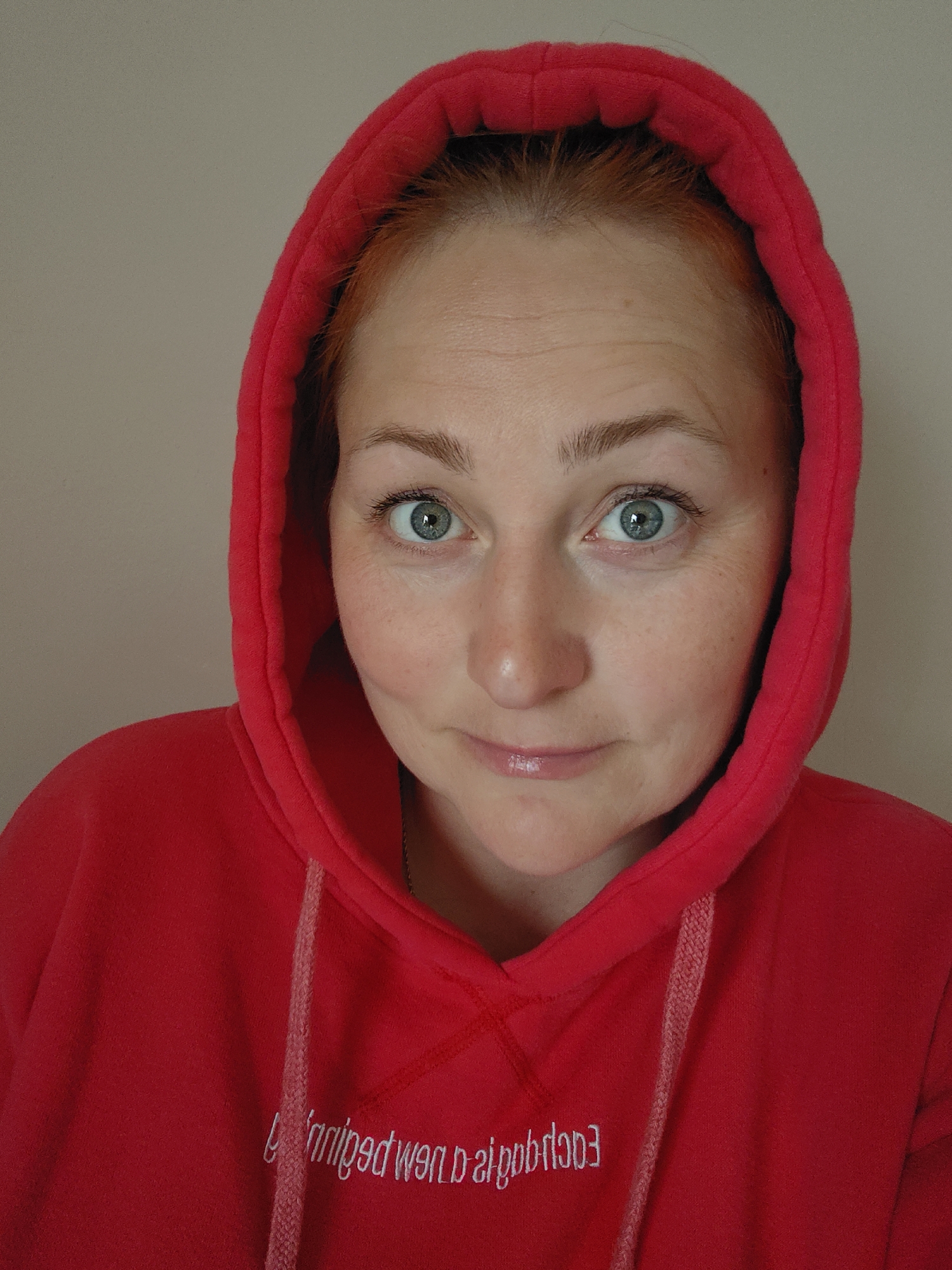 A close-up portrait shows a woman looking directly at the camera, filling most of the frame from the chest up. She has fair skin, light red or auburn hair visible at her hairline, and striking blue-green eyes that are wide and alert. Her eyebrows are light brown and well-defined. Faint lines are visible on her forehead and around her eyes, and she has a scattering of light freckles across her nose and cheeks. Her lips are slightly parted in a gentle smile and appear glossy.
She is wearing a vibrant red hooded sweatshirt (hoodie) with the hood up, framing her face. The fabric appears soft, possibly fleece. A light-colored, braided drawstring hangs down the front of the hoodie. White embroidered text is visible on the front of the sweatshirt, appearing mirrored in the image. When corrected, the text reads "The new beginning." The background is a plain, light, neutral color, suggesting an indoor setting in Markovskii, Russia. The lighting is even, highlighting her features and the texture of her clothing. The overall impression is one of candidness and a subtle, inquisitive expression.