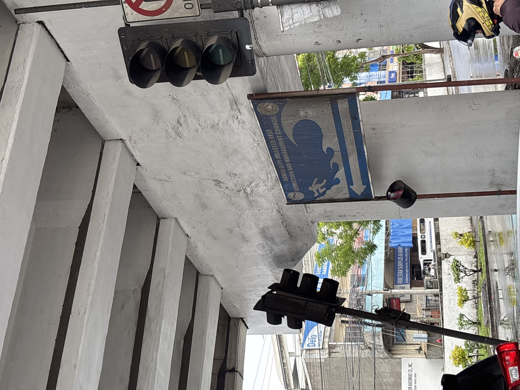 A traffic light hangs angled from a pole beneath an overpass in Davao City, Philippines. To the right, another traffic light is mounted on a pillar. A blue and white sign displaying a wave and a person running is also attached to the pillar. Buildings and trees are visible in the background, with a wet road suggesting recent rain.