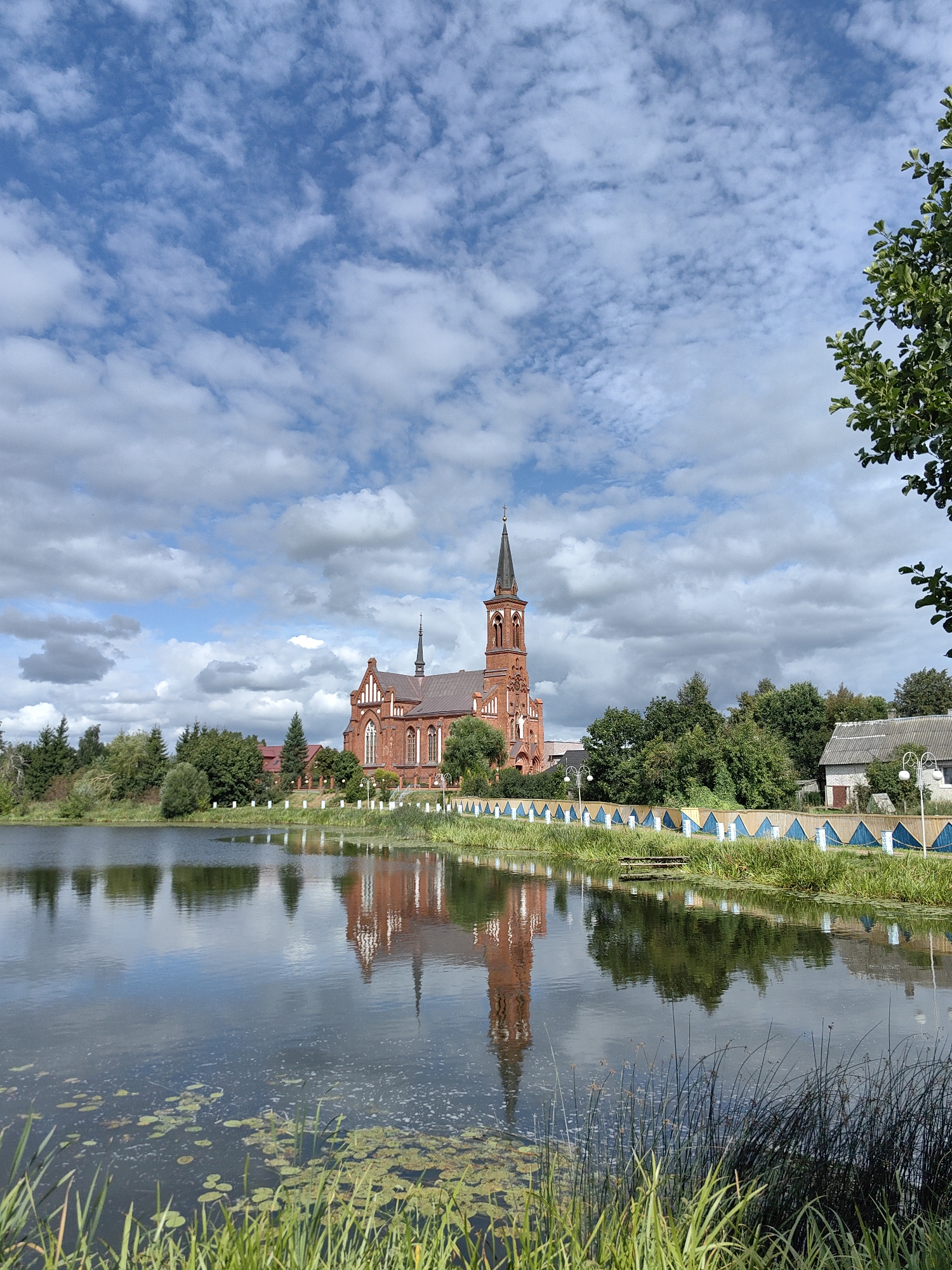 The image depicts a picturesque view of a red brick church with a tall spire, reflected in a calm lake. The church is situated across the lake, surrounded by lush green trees. A white fence with blue accents runs along the lake's edge in front of the church. The sky is a vibrant blue with scattered white clouds. The foreground of the image features tall green grass and water lilies on the surface of the lake. The scene is idyllic and serene, suggesting a peaceful atmosphere. The location is the Church of the Exaltation of the Holy Cross in Pastavy, Belarus.