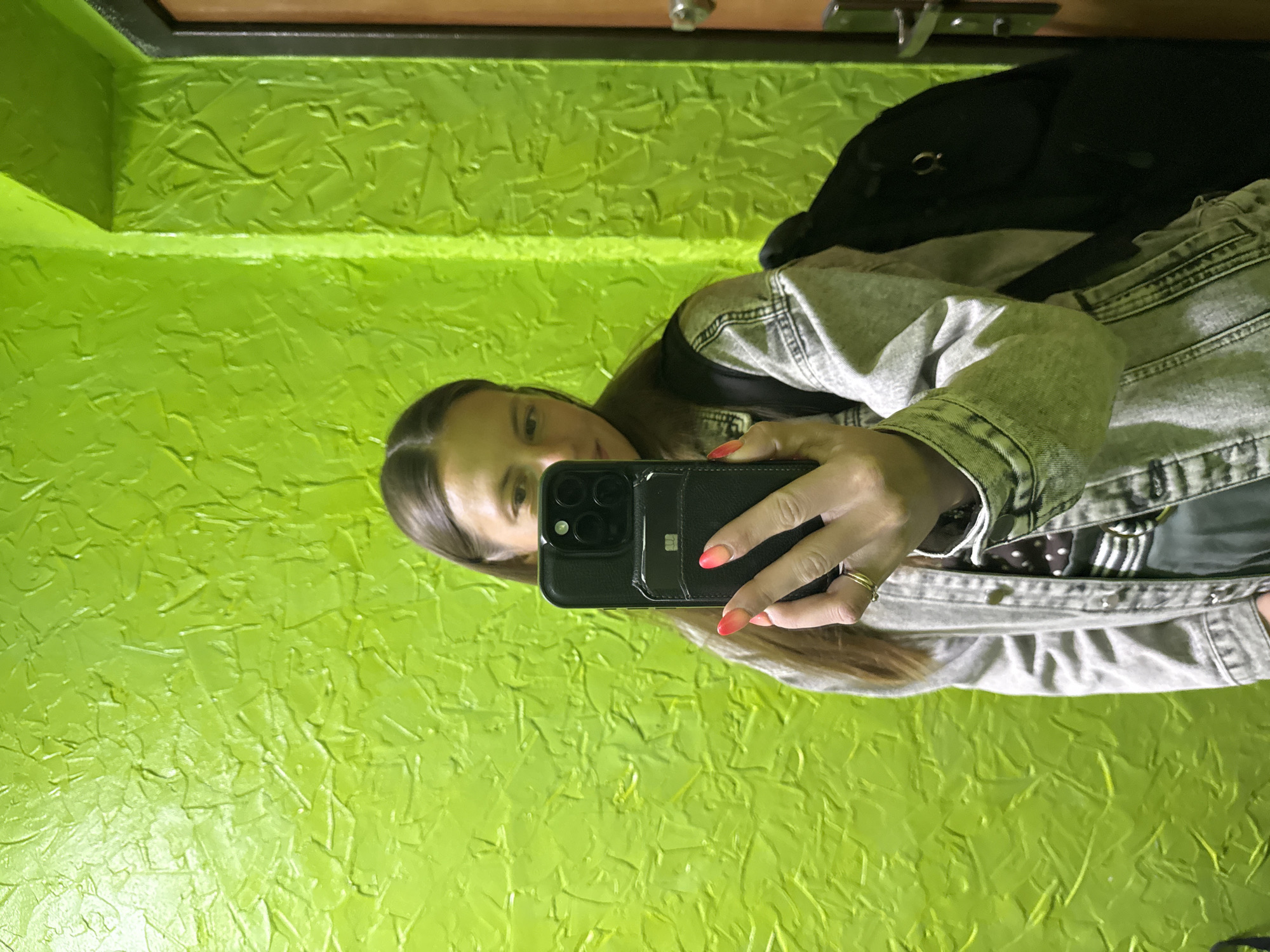 A person with long, dark hair is holding a black smartphone in their right hand, taking a selfie. They are wearing a light gray denim jacket over a black-and-white polka-dot shirt. A black backpack is visible behind the person. The background is a bright green wall with a textured, stucco-like finish. Above the wall, a brown surface with metal hardware is visible. The image appears to be taken in an indoor setting. It is not possible to verify that the location of this image is Chaikovsky, Russia.