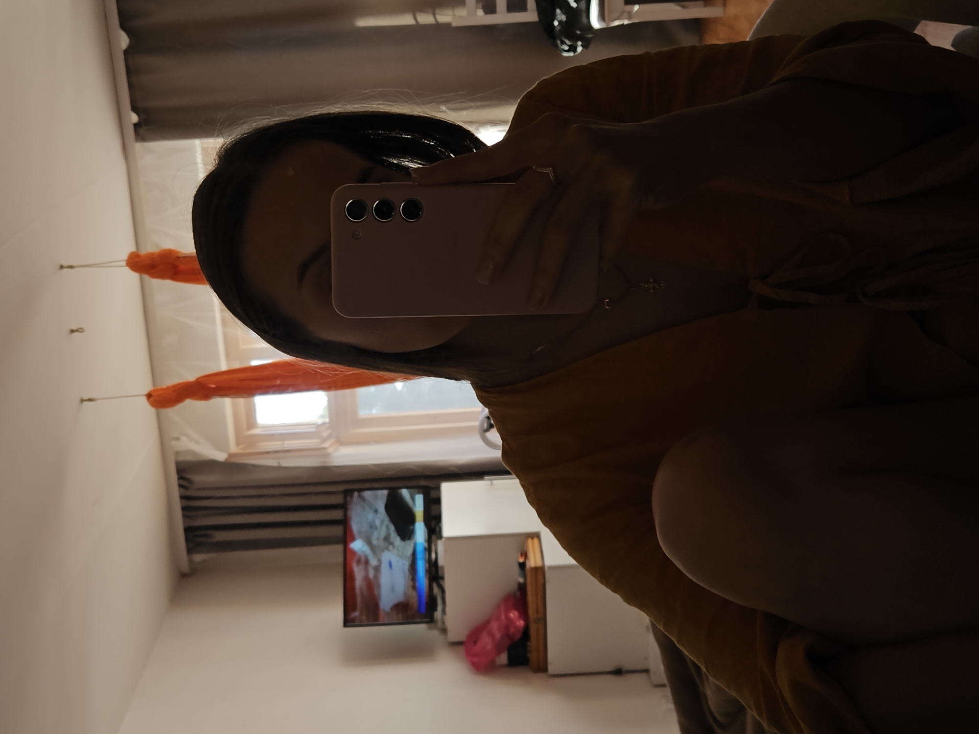 In an indoor setting, a person takes a mirror selfie using a pink smartphone. They are wearing a yellow garment and have a necklace with a pendant. The room appears to be a bedroom or living area, with a television set on a white cabinet in the background. Orange fabric hangs from the ceiling. The lighting suggests it is daytime, possibly late afternoon or evening, with natural light filtering through a window. The exact location in Chaikovsky, Russia, cannot be determined from the visual information provided.