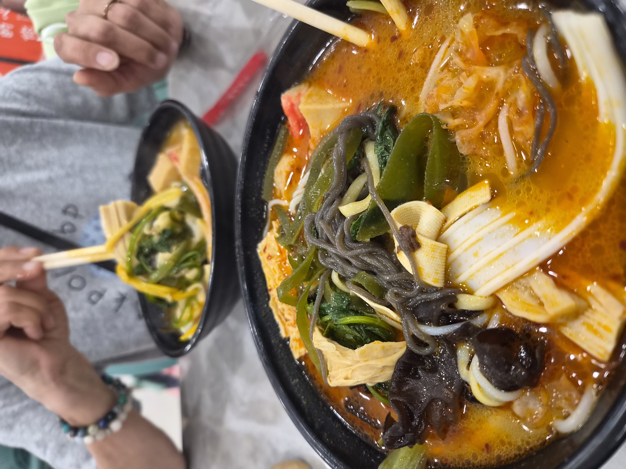 A close-up shot shows two bowls of hot pot at a table in Mishan, China. The bowl in the foreground is filled with a red broth, noodles, leafy green vegetables, dried tofu, and dark mushrooms. Chopsticks are visible in the bowl. In the background, another person is picking up noodles from a second bowl with chopsticks. They are wearing a gray t-shirt and a bracelet on their left wrist. The table is gray and speckled.