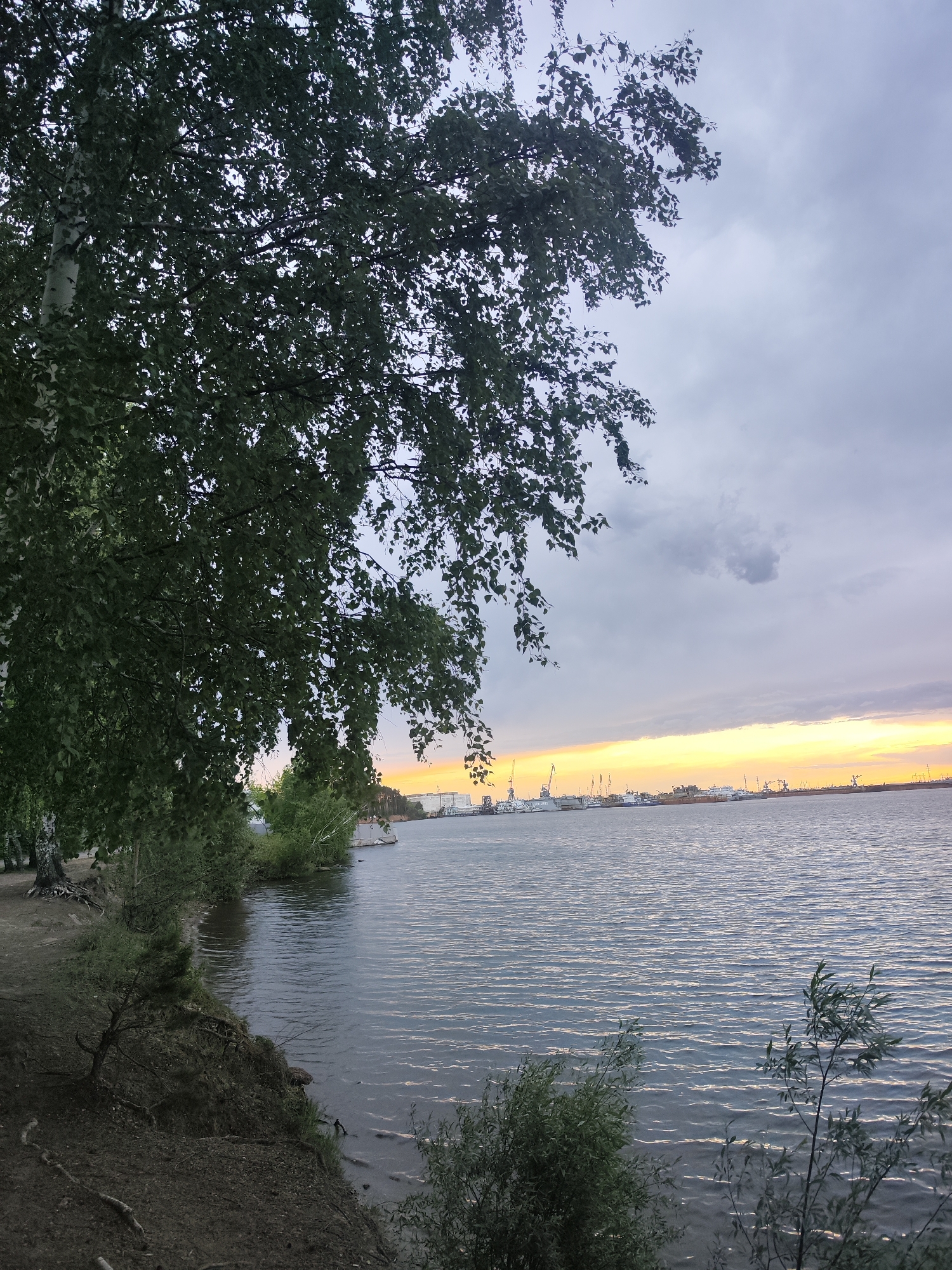 The image shows a view of the Kama River in Chaikovsky, Russia, at twilight. The foreground features the riverbank with trees and foliage. The river stretches towards the horizon, reflecting the orange and yellow hues of the setting sun. In the distance, a silhouette of industrial structures, including cranes and ships, lines the far shore. The sky is overcast with gray clouds. There are no people visible in the image.