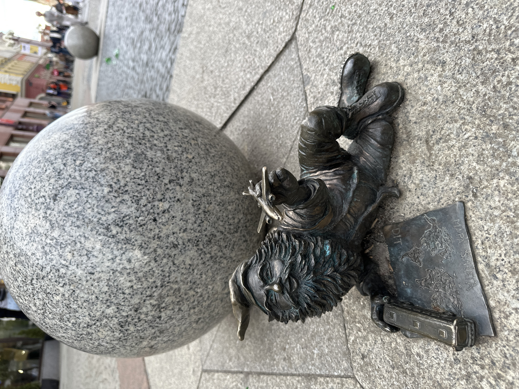 The image depicts a bronze dwarf statue in Wrocław, Poland, resting on a gray stone-tiled ground next to a large, similarly colored granite sphere. The dwarf is lying on its side, its legs crossed at the ankles. It has a beard and wears what appears to be a tunic. In its hands, it holds a pair of scissors and a metal sheet with the words "DIT MAPA SWIATA" (possibly a distorted "DIT MAPA ŚWIATA," meaning "DIT World Map") and an image resembling a world map, along with what looks like a miniature train car. The background shows a blurred street scene with pedestrians and buildings. The statue is one of the city's famous dwarves that are scattered throughout Wrocław.