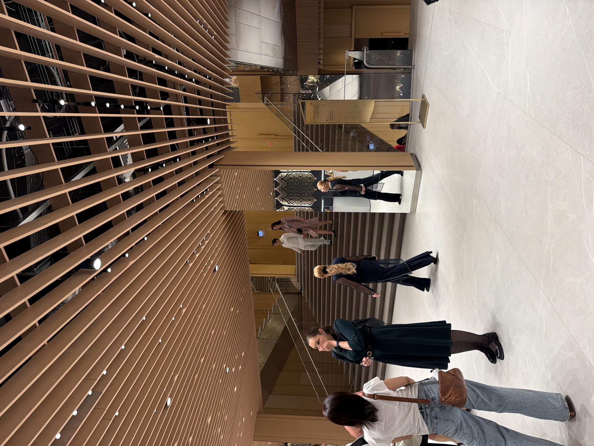 A group of people are gathered in a modern, indoor architectural space. The scene is dominated by a slatted wooden ceiling that extends from the left side of the frame to the top right, with a series of small, circular lights embedded within the slats. The floor is tiled with large, light-colored tiles that have subtle white veining.
In the mid-ground, a set of stairs with a glass railing is visible, leading upwards. Several individuals are navigating this space. On the stairs, a woman with blonde hair wears a dark sheer dress. Below her, on the lower level of the stairs, a woman in a dark green dress with black tights and shoes is looking downwards, holding her hand to her mouth. Further down, a person in a white top and blue jeans is seen from the back, carrying a brown shoulder bag.
In the background, a person in a patterned dress and another in a white outfit are walking up the stairs. A decorative metal screen with an intricate pattern is mounted on a wall. Mirrors are strategically placed, reflecting parts of the scene. The lighting suggests it is daytime, and the environment appears to be a public building such as a museum, gallery, or a modern commercial space. There are no visible signs of the current weather conditions. The overall impression is one of a leisurely visit or a gathering.