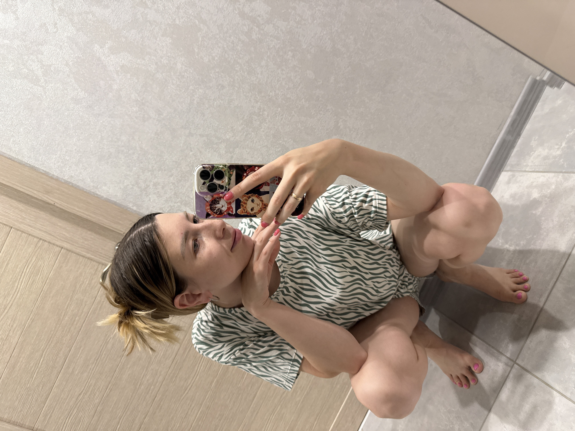 A young woman is squatting in a bathroom, taking a mirror selfie. She is wearing a green and white striped t-shirt and has her hair pulled up in a messy bun. Her fingernails are painted pink, and she is wearing a ring on her left hand. The bathroom has beige tiled walls and a wooden door. The floor is tiled in gray. A phone with a cat-themed case is visible in her right hand. The image is taken from a high angle, looking down at the woman. There is no visible indication of the location being Moscow, Russia.