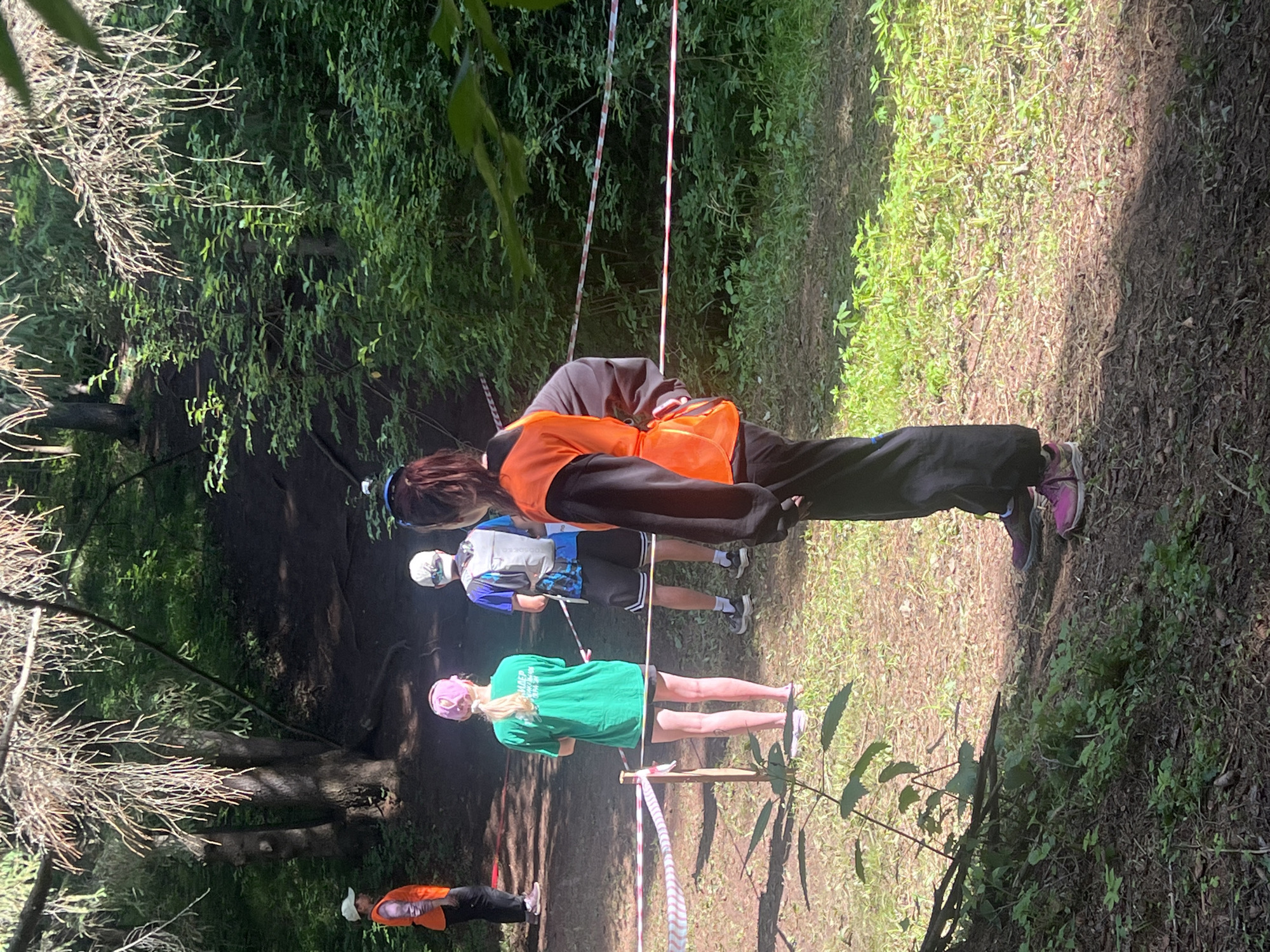 The image shows a group of people standing in a wooded area, possibly a park or forest. A red and white striped tape is stretched across the scene, suggesting a boundary or a race course. The people are dressed in casual clothing, and some are wearing vests. One person is wearing an orange vest, and another appears to be wearing a blue and white vest. The environment is characterized by trees, lush green foliage, and a dirt path. The lighting suggests it is daytime, with sunlight filtering through the trees. The overall scene appears to be related to an outdoor activity or event. The location is stated to be in the city of Сергино, Russia.