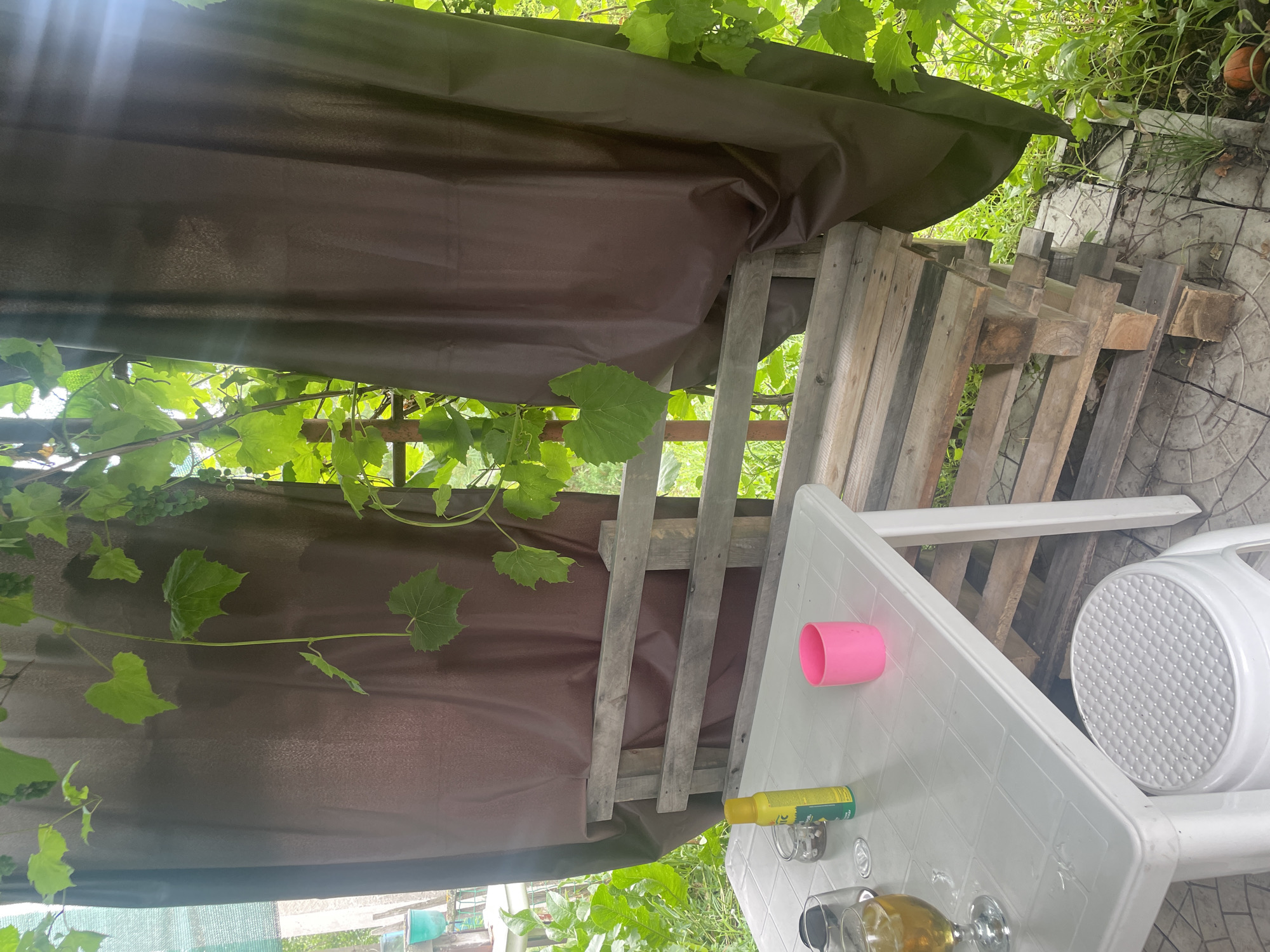 The image depicts an outdoor seating area, possibly a patio or garden space, in a rustic setting. A white plastic table is positioned on the left, with a glass of amber liquid, a small jar, a spray can, and a pink cup on its surface. Beneath the table, a white plastic stool with a textured seat is visible. In the background, a bench constructed from wooden pallets is situated. The entire scene is partially covered by dark brown fabric curtains, with lush green grapevines with large leaves and small clusters of green grapes hanging overhead and to the sides, suggesting a warm, possibly summer, environment. The ground appears to be paved with circular tiles. The overall impression is of an informal, DIY outdoor relaxation spot.