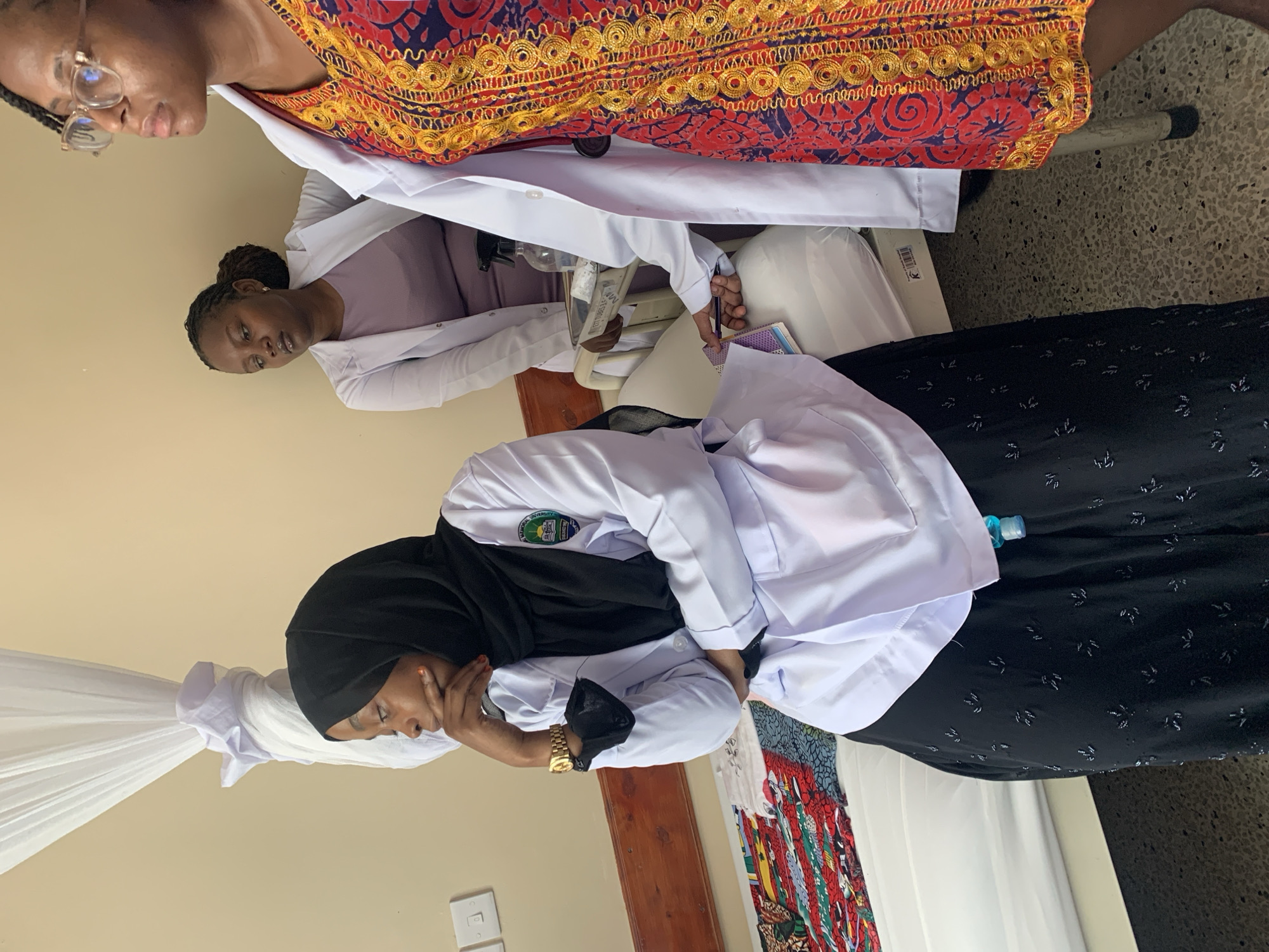 Three individuals are present. One woman wears a red and gold patterned dress, glasses, and a white coat. Another woman wears a white coat over a purple shirt, and the third woman wears a black hijab and a white coat. The woman in the hijab sits with her head in her hand. The two other women are standing and writing on medical charts. The location appears to be a medical facility. It is difficult to ascertain whether the image was taken in Dar es-Salaam, Tanzania.