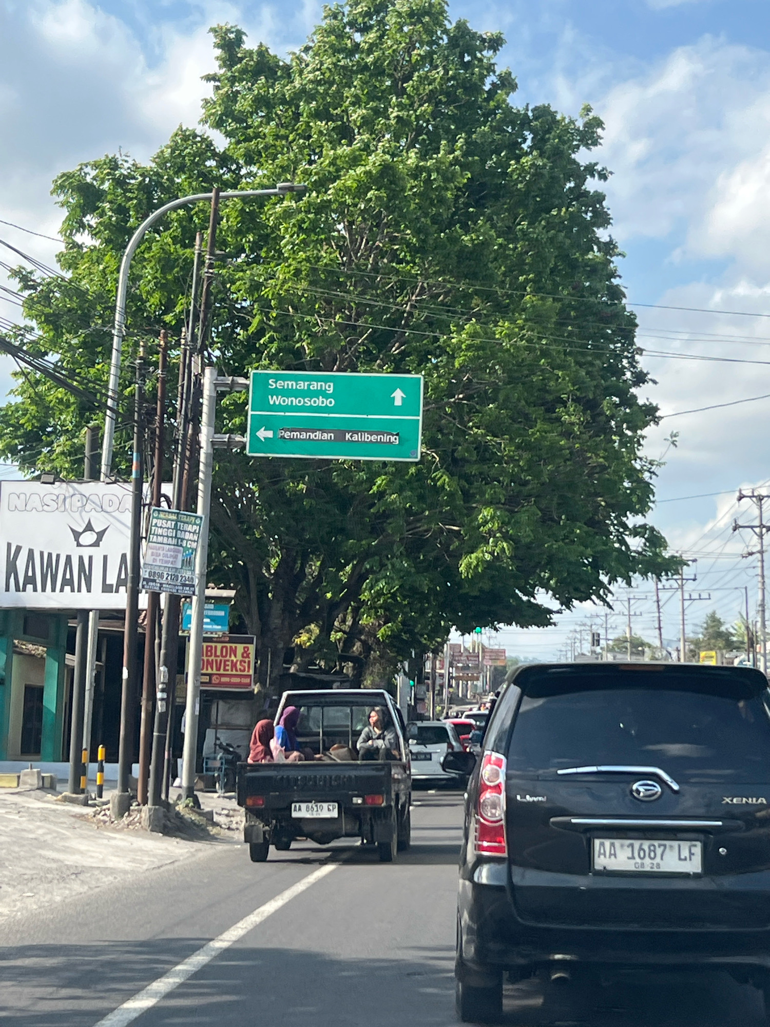 The image depicts a street scene in Payaman, Indonesia. A large green directional sign indicates routes to "Semarang Wonosobo" and "Pemandian Kalibening". In the foreground, traffic is moving along a road. A black pickup truck with three passengers is in the left lane. The passengers are women wearing headscarves. In the right lane, a black SUV with a license plate reading "AA 1687 LF" is visible. The surrounding environment includes a large tree with lush green foliage, utility poles with numerous wires, and buildings on the left side of the road. The sky is partly cloudy.