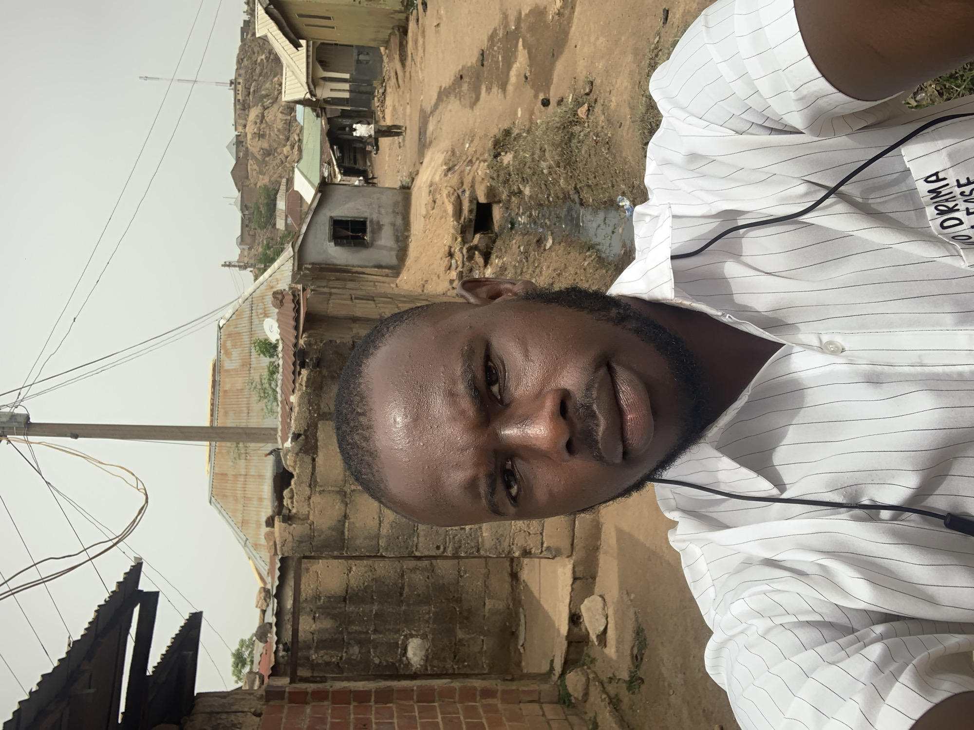 A medium, close-up shot shows a man in a striped white shirt. He has dark hair and a beard. The location appears to be in Jos, Nigeria. The background reveals what seems to be a poor neighborhood with dirt streets and simple, rough-looking buildings. A rocky hill is visible in the distance. A power pole is visible.