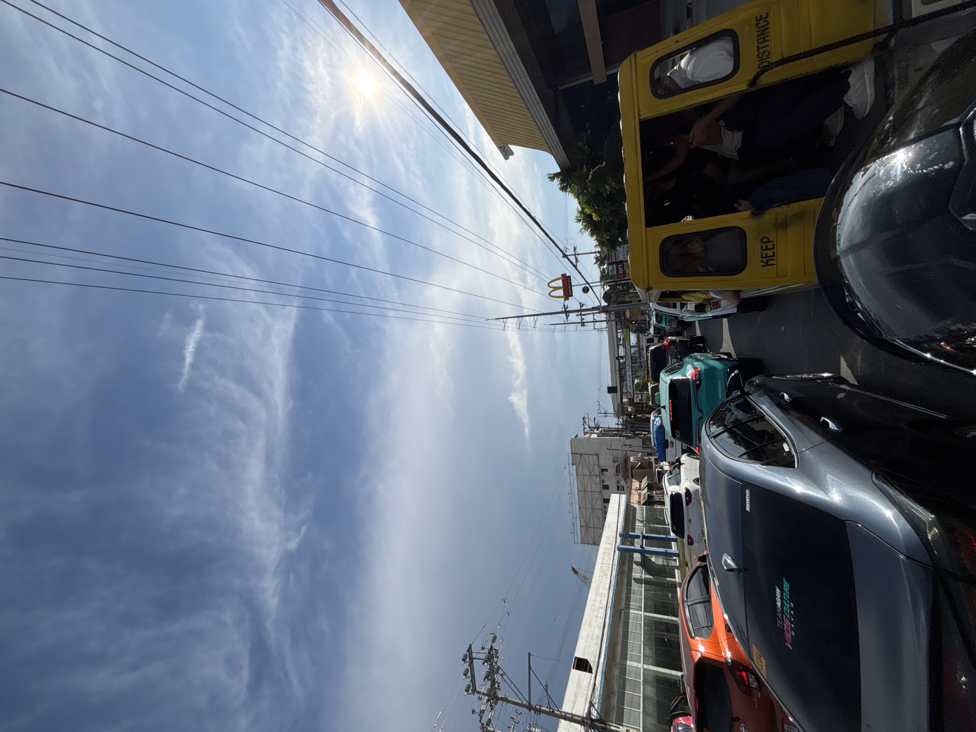The image captures a street scene in Davao City, Philippines, likely during daytime, as evidenced by the bright sun and clear sky. The perspective is from within a vehicle, looking forward and slightly upwards. A traffic jam is evident, with numerous cars and a yellow multicab filling the road. The multicab, a common mode of transport in the Philippines, has people inside and a "KEEP DISTANCE" sticker on its side. A McDonald's sign is visible in the background, suggesting a commercial area. Utility poles and power lines crisscross the sky, a common sight in urban Philippine landscapes. The sky is a mix of blue with wispy white clouds, with the sun shining brightly, creating lens flare. The buildings lining the street appear to be commercial establishments. The overall impression is a busy urban environment with characteristic Filipino street life.