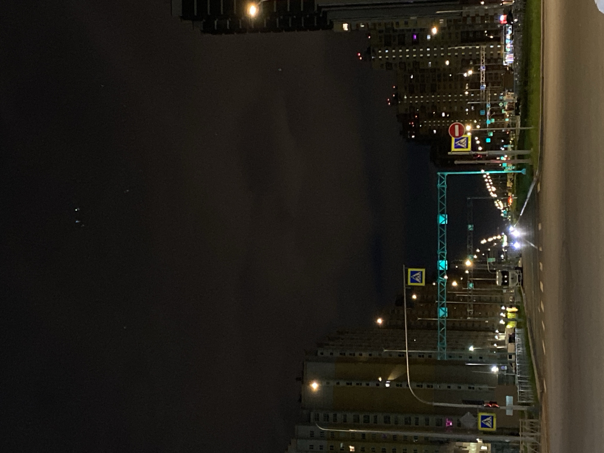 The image depicts an urban night scene in Saint Petersburg, Russia. The perspective is from a vehicle moving along a multi-lane road. Tall apartment buildings line both sides of the street. Streetlights illuminate the road and surroundings. Road signs, including pedestrian crossing and no entry symbols, are visible. An overhead gantry-like structure spans the road. The sky is dark. No people are discernible. A truck is visible on the road ahead.