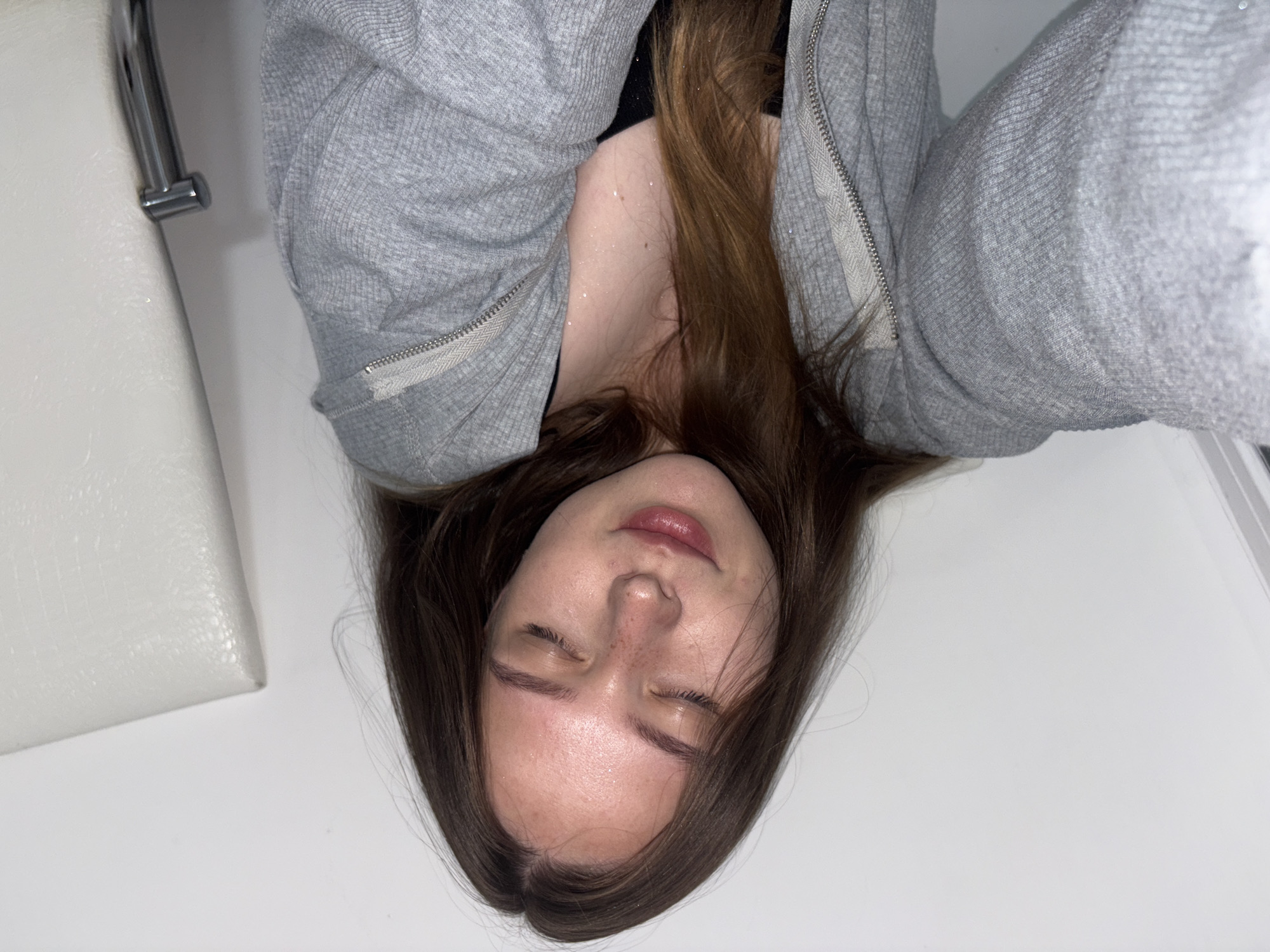 The image shows a close-up of a young woman with her eyes closed, resting her head against a white surface. She has long, brown hair and freckles on her nose and cheeks. She is wearing a gray zippered hoodie, and a hint of a black bra is visible. There are small, sparkly particles on her neck and chest, which may be glitter or sweat. The background is a plain white wall, and a metallic faucet is visible in the upper right corner, suggesting the image was taken in a bathroom. The photograph is taken from a slightly tilted angle. The location of Chaikovsky, Russia, is not discernible from the image itself; it is an external detail provided as context.