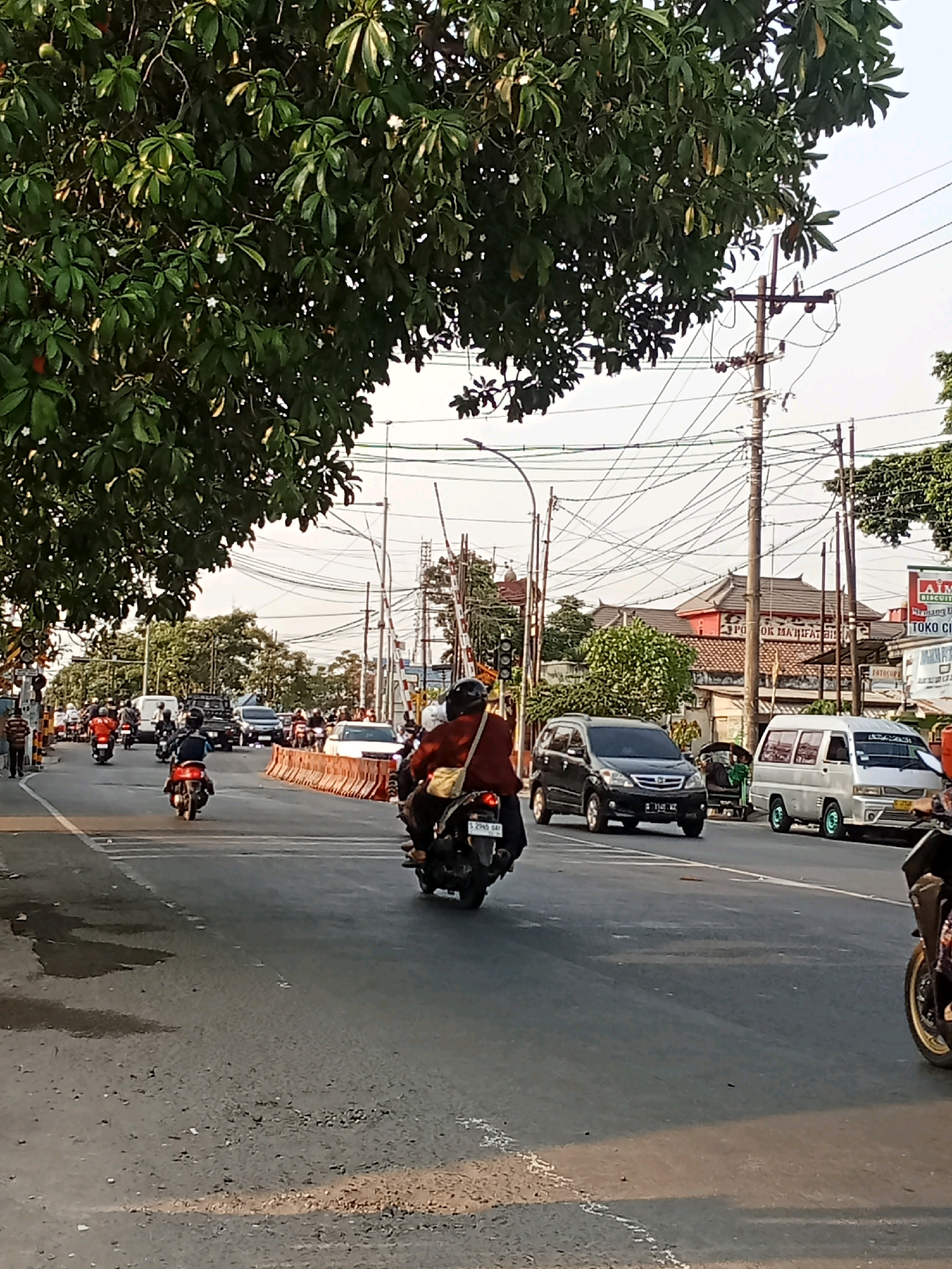 The image depicts a street scene in Kaliwungu, Indonesia, under a bright sky, possibly late afternoon given the lighting. A busy road is visible with several vehicles, including motorcycles, cars, and vans. In the foreground, a person on a motorcycle is traveling away from the camera. Other motorcycles are also present on the road. On the right side of the road, a white van is parked, and further down, a silver van is moving. A black car is also seen in traffic.
The environment features abundant trees with green foliage, particularly on the left side of the image, casting shadows on the road. Utility poles with a dense network of overhead wires are prominent throughout the scene, indicating an urban or semi-urban setting. Buildings are visible in the background, some with traditional Indonesian architecture featuring red accents and tiled roofs. Traffic control barriers, orange in color, are placed on the road, suggesting a possible road closure or diversion. A traffic light signal is visible on the left side of the road. Advertisements and signage are present on some of the buildings. The road surface appears to be asphalt, with some visible wear and tear and scattered debris. The overall impression is of a dynamic, everyday street environment in Indonesia.