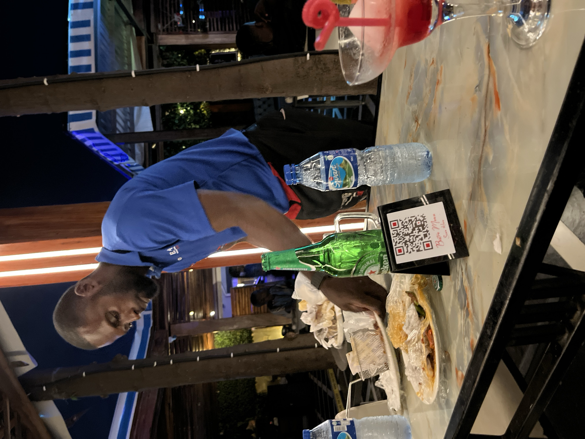 A Black man in a blue polo shirt and black pants is standing behind a table laden with food and drinks at an outdoor establishment. He appears to be a server or staff member, as he is reaching for a plate of food. On the table are two bottles of water, one empty and one partially full, a green bottle of Heineken beer, and several plates of food, some covered in plastic wrap, suggesting that a meal has recently been consumed. A QR code stand with the text "Bistro Menu" is also visible. To the right, a margarita glass with a pink drink and a straw is positioned on the table. The background shows a dimly lit outdoor seating area with wooden beams, striped awnings, and distant lighting. This scene likely takes place at a restaurant or bar in Abuja, Nigeria, given the visual context.
