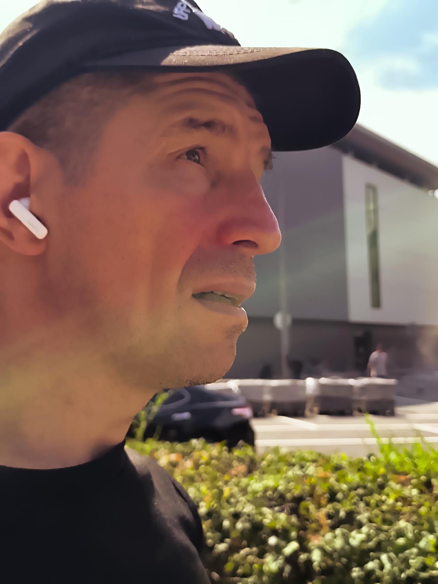 A man wearing a black baseball cap and a black t-shirt is visible in a close-up, side-profile shot. He has an earbud in his left ear. He appears to be outdoors in a city setting, with a modern building and a blurry street scene in the background. The lighting suggests it is daytime. The man's facial expression is neutral. The specific location within the city administration of Qarağandı, Kazakhstan, is not discernible from the visual information in the image.