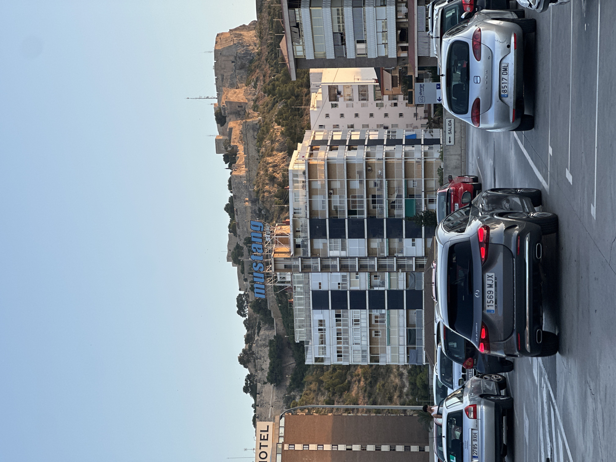 The image depicts a cityscape with a large fortress on a hill in the background. In the mid-ground, there are several multi-story buildings, some with apartments and balconies, and one with a large "mustang" sign. In the foreground, there is a parking lot with several cars. One silver car in the foreground has a license plate that reads "1569 MJX." Another silver car to the left has a license plate that reads "2785 BXH." A red car is parked behind the silver car with license plate "2785 BXH." A sign with the word "SALIDA" (Spanish for "EXIT") is visible on the right side of the parking lot. The sky is clear and blue, suggesting it is daytime, possibly late afternoon given the lighting on the fortress.