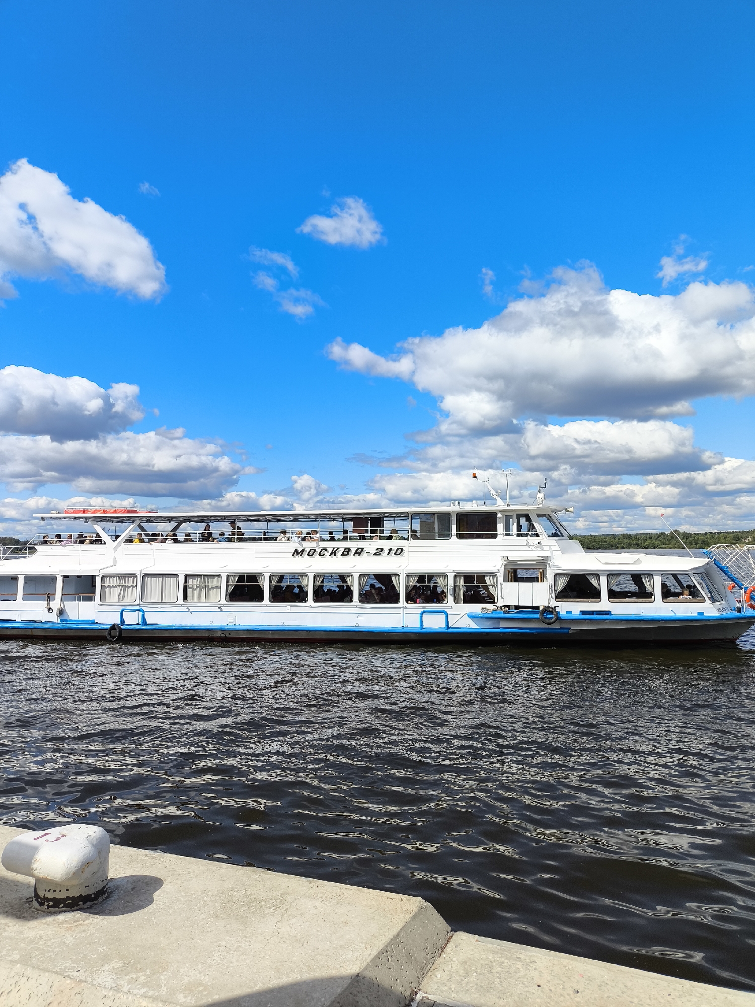 A white passenger boat with a blue stripe is docked on a dark body of water, likely the Kama River, in Perm, Russia. The boat, named "МОСКВА-210," has multiple windows and a second deck with passengers visible. The sky is bright blue with scattered white clouds. In the foreground, a concrete pier with a white mooring bollard is visible. The overall scene depicts a sunny day with a boat ready for or returning from a journey.