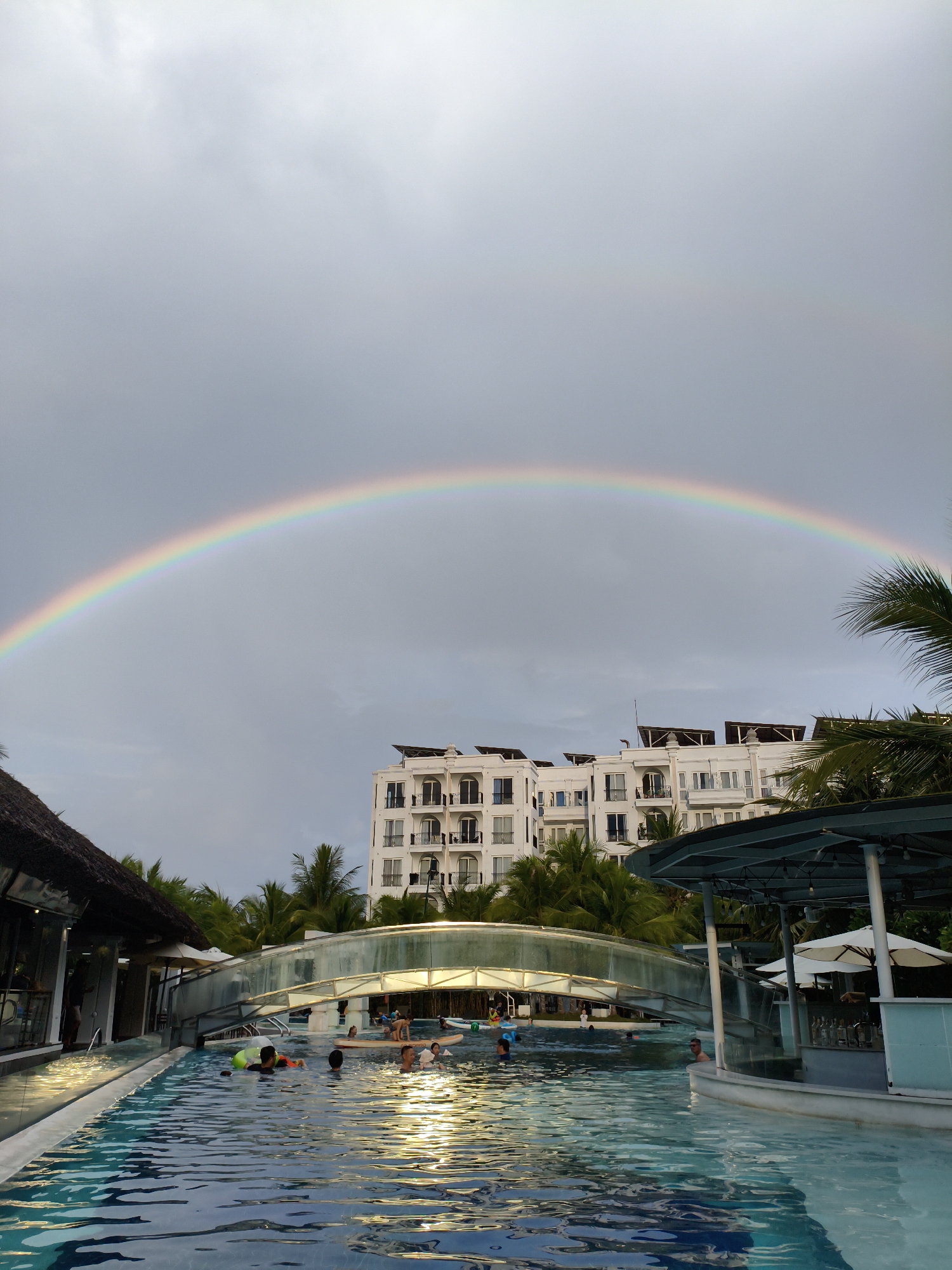 The image depicts a resort swimming pool with a rainbow arched across the sky. In the background, a multi-story white building with balconies and solar panels is visible, along with palm trees. The pool itself is filled with water, reflecting the sky and surrounding lights. Several people are in the pool, some swimming and others interacting, with some children visible. A bridge spans over a section of the pool, and a bar area with white umbrellas is to the right. The scene suggests a tropical resort setting, likely in Nha Trang, Vietnam, given the architectural style and vegetation. The sky is overcast with a rainbow prominently displayed.