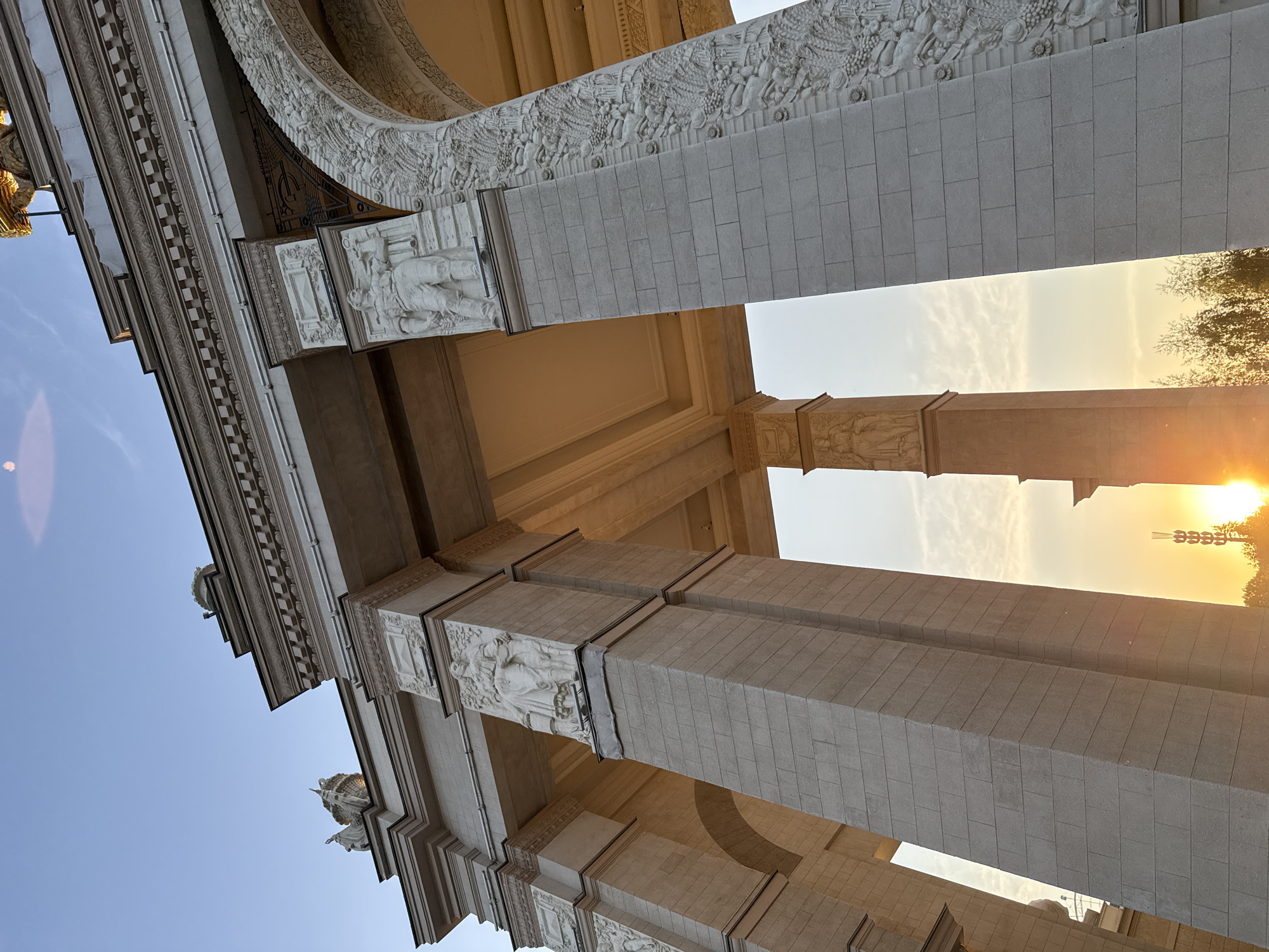 The image shows a low-angle view of the Main Entrance Arch of the VDNKh (Exhibition of Achievements of National Economy) in Moscow, Russia. The arch is composed of large, stone-like blocks with relief carvings and sculptures. The sky is visible through the arch, with sunlight breaking through on the right side of the frame. The building appears to be under the warm light of either sunrise or sunset. The architectural structure consists of grand arches with intricate detailing and classical ornamentation.