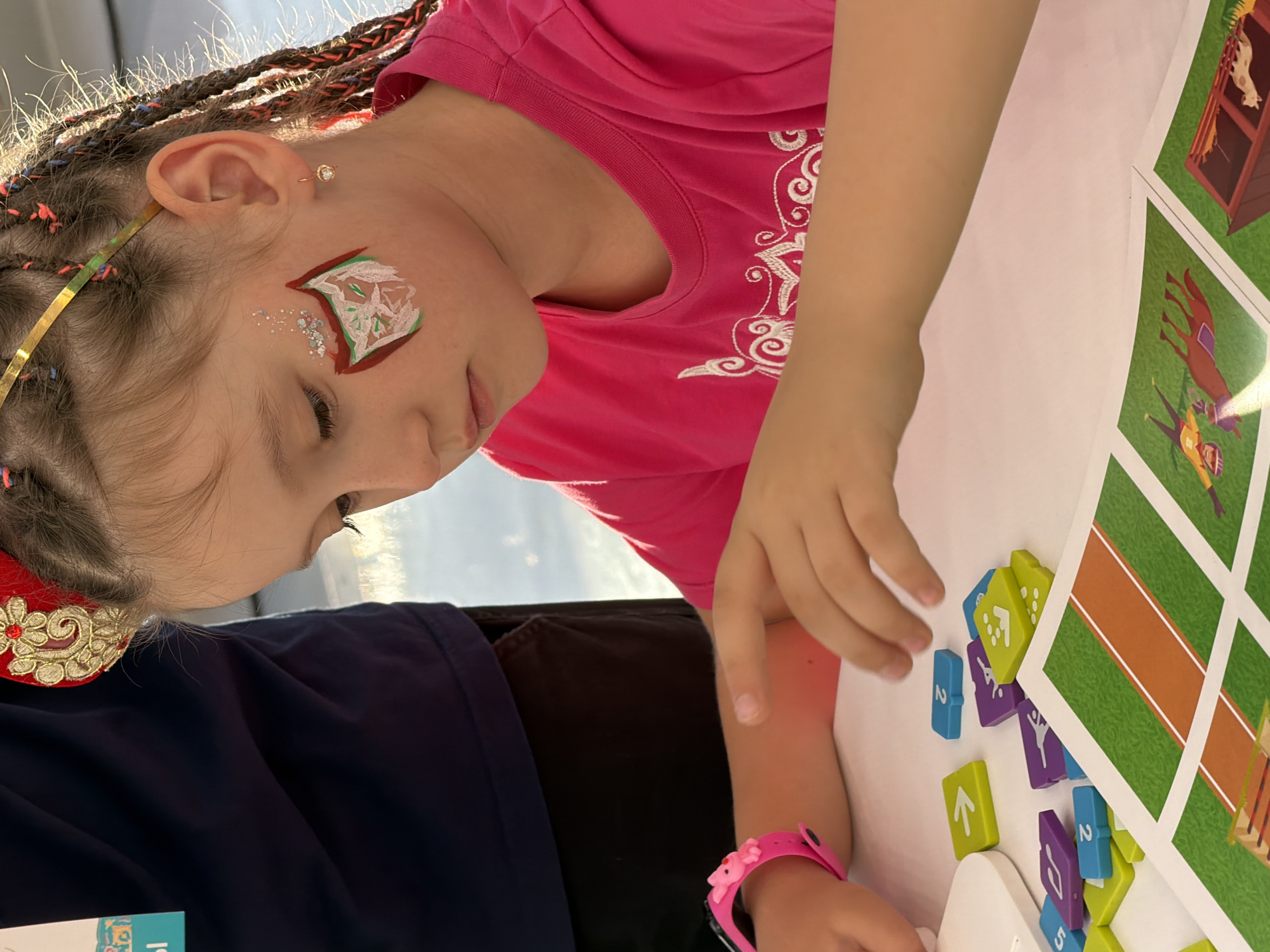 A young girl with face paint on her left cheek and braids in her hair sits at a white table, looking down at a game. She is wearing a bright pink t-shirt with white embellishments. In front of her are colorful game pieces, mostly green and purple, with numbers or icons on them, as well as game boards with illustrations of a racetrack and farm animals. A person in a dark blue shirt is visible to the left of the frame, partially obscuring the girl. The lighting suggests an indoor setting with sunlight coming from the right.