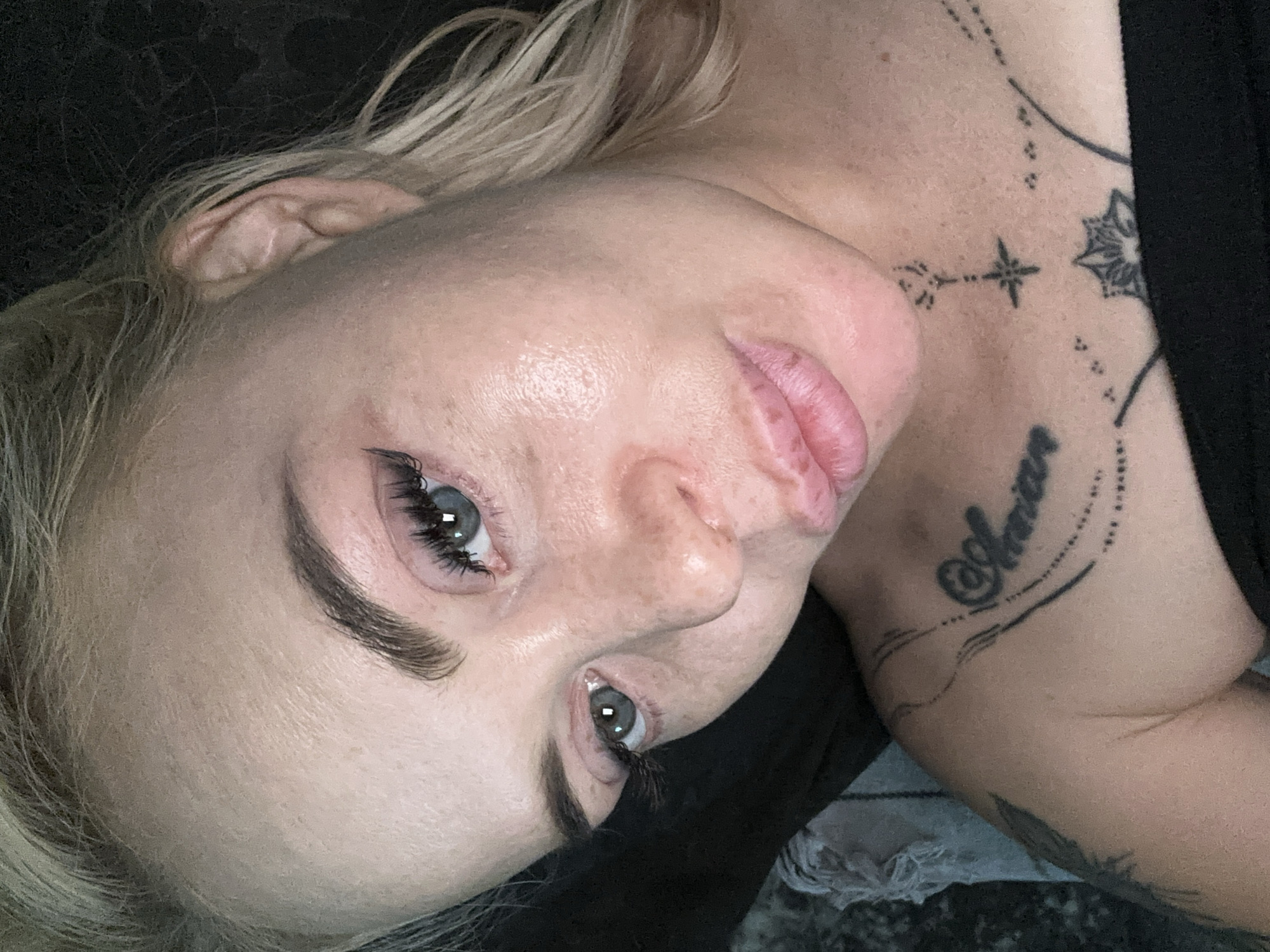 A close-up, high-angle selfie shows an adult female with fair skin, light blonde hair featuring darker roots, and light blue-grey eyes, located in the city of Unknown, Unknown. She looks directly into the camera with a slight head tilt, her full, pink lips parted in a soft pout. Her eyebrows are dark and defined, accentuated by long, dark eyelashes and black winged eyeliner. Her facial skin displays visible pores and minor texture variations, with a localized sheen on her forehead and cheekbones. Several black-ink tattoos are visible: a script design with decorative elements on her left collarbone, and a small compass rose/star positioned centrally on her upper chest with a lotus-like floral pattern below it. A black strapless garment covers her chest. The background is dark and largely indistinct, with a light grey, textured object partially visible to the left of her shoulder. The scene is illuminated by direct lighting, creating subtle shadows.