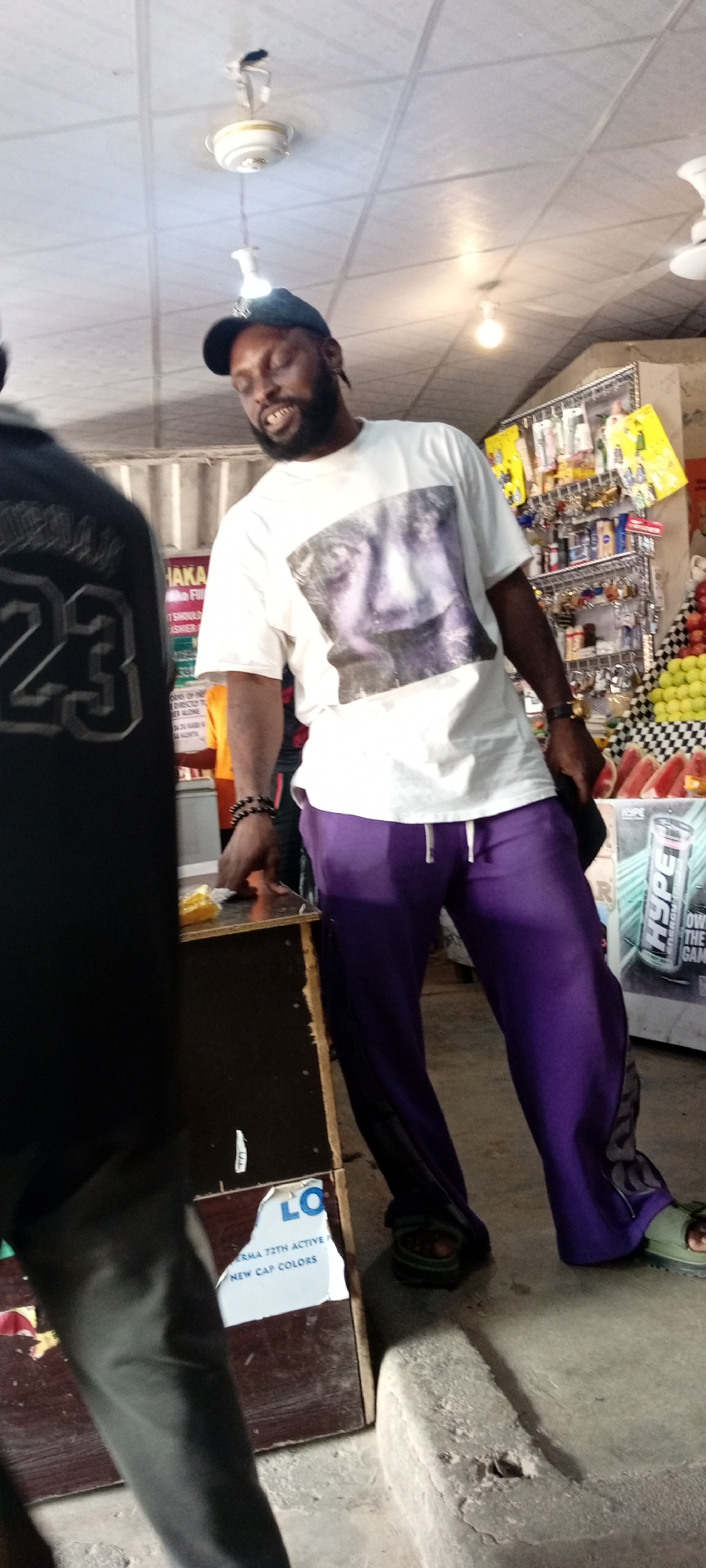 The image shows a man wearing a white t-shirt with a large black and white portrait on it, a dark blue baseball cap, and bright purple sweatpants. He is standing in what appears to be a small shop or market stall, with shelves stocked with various items behind him. To his left, another person is partially visible, wearing a black shirt with the number "23" on the back. The man in the foreground is looking down and to his right, with his mouth slightly open, and he is holding a dark object in his right hand. He has a beard and is wearing bracelets on his left wrist. The setting is indoors, with a ceiling fan and fluorescent lights visible. A display of sliced watermelon and other fruits can be seen to the right of the man. There is also text visible on posters and packaging in the background, including "HYPE ENERGY DRINK" and "NEW CAP COLORS". The overall impression is a candid shot of a man in a bustling commercial environment in Aviation Quarters, Nigeria. The lighting suggests it could be daytime, though the indoor setting makes it hard to determine.