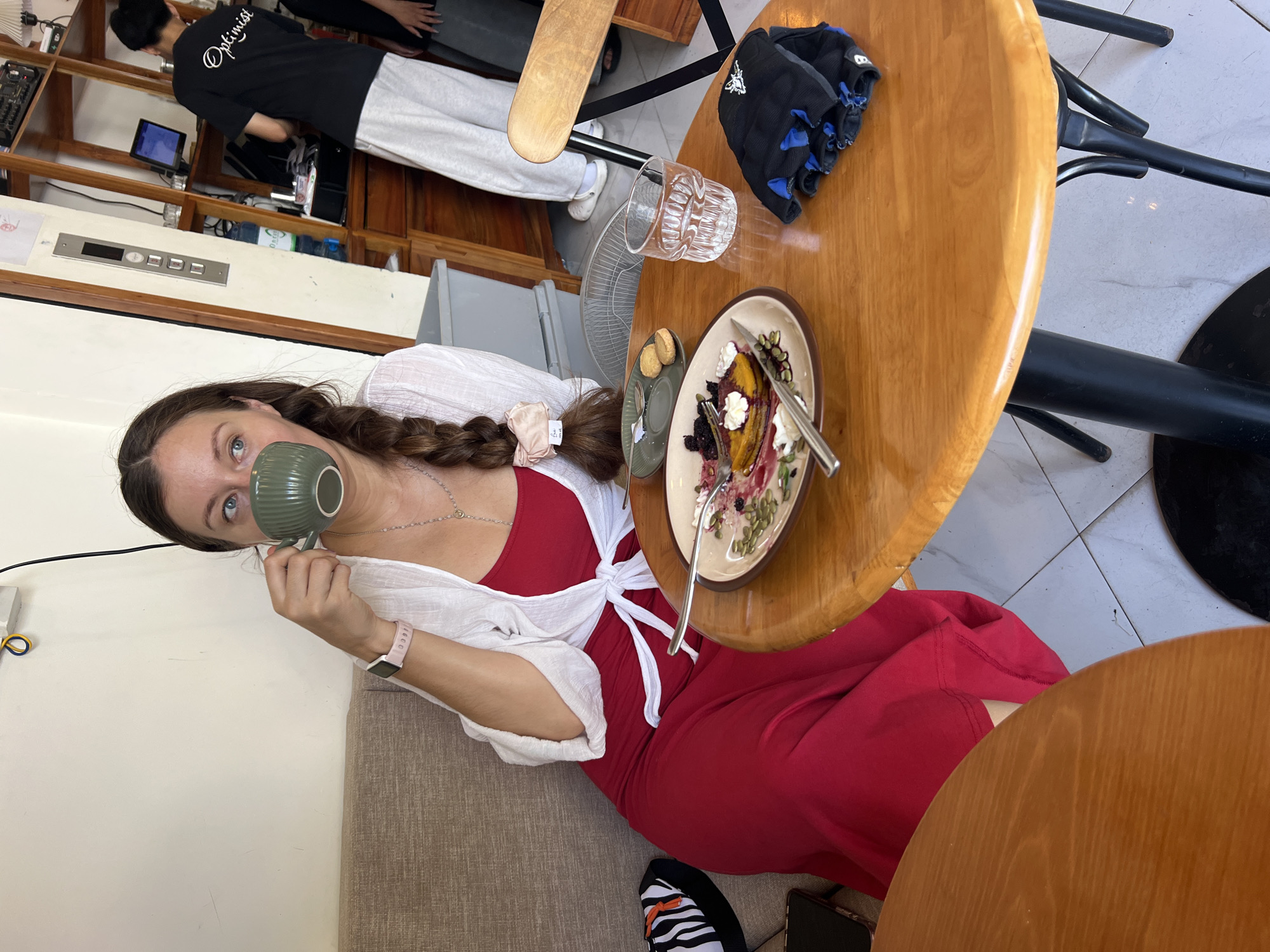A woman is sitting on a couch in what appears to be a café in Nha Trang Ward, Vietnam, as suggested by the Vietnamese text on the menu in the background. She is drinking from a green cup. She wears a red dress, a white cardigan tied at the waist, and an Apple Watch. Her hair is styled in a braid.
On the table beside her are a plate of food with a slice of cake, a glass of water, a small plate with cookies, and black gloves. Other patrons are in the background. The café has wooden tables and chairs, along with tiled flooring.