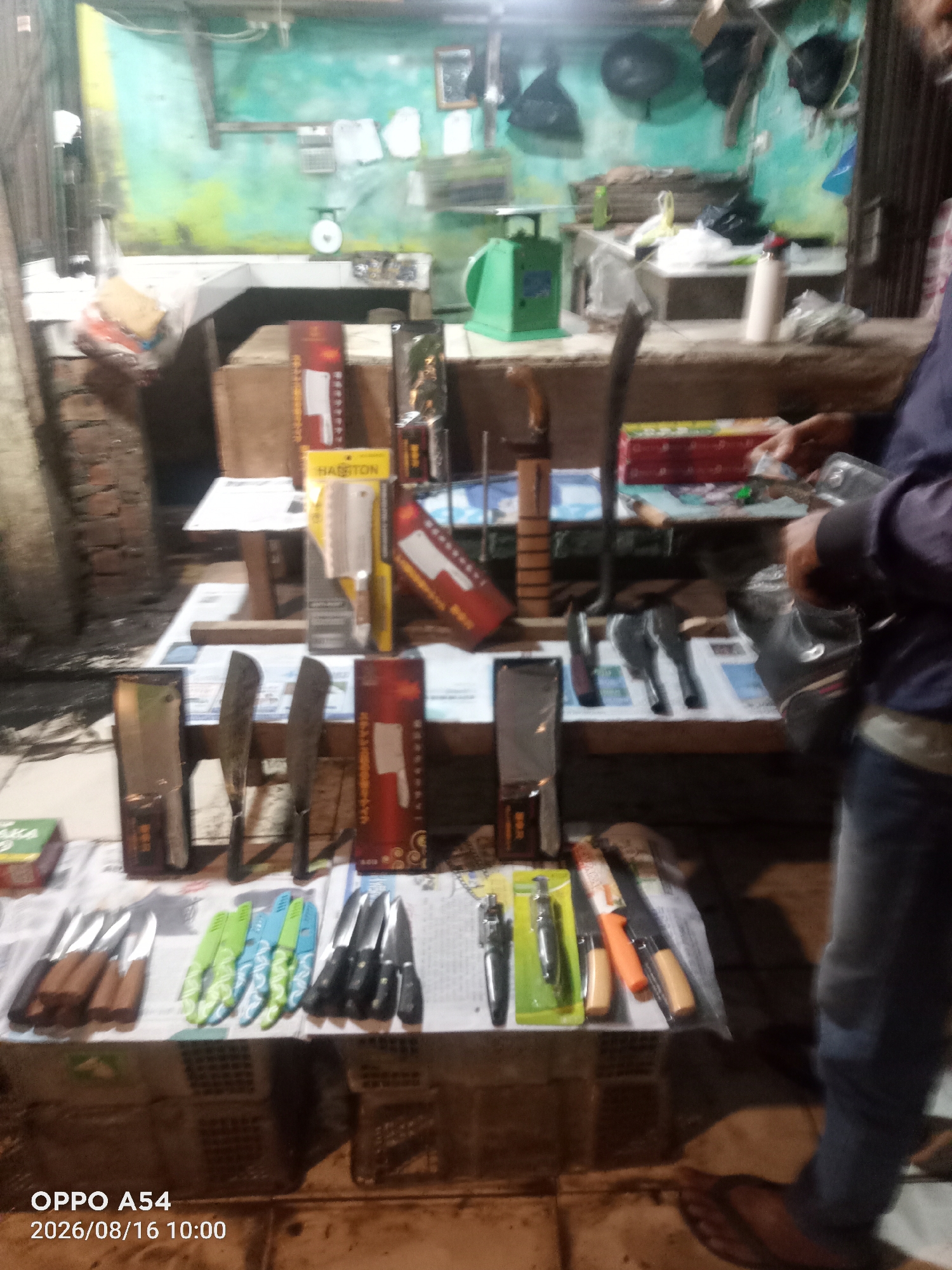 The image depicts a stall selling kitchen knives and related tools, likely in a market or roadside setting in Jambi, Indonesia. The stall is organized with various knives displayed on a table, some in packaging and others loose. In the background, there appears to be a counter with items like a scale, green containers, and other unidentified objects. A person, only partially visible from the waist down, is standing to the right of the stall, wearing blue jeans and sandals, and appears to be interacting with the items on display. The lighting suggests it might be evening or a dimly lit area. The overall scene is cluttered but organized, indicating a commercial transaction or display of goods.
