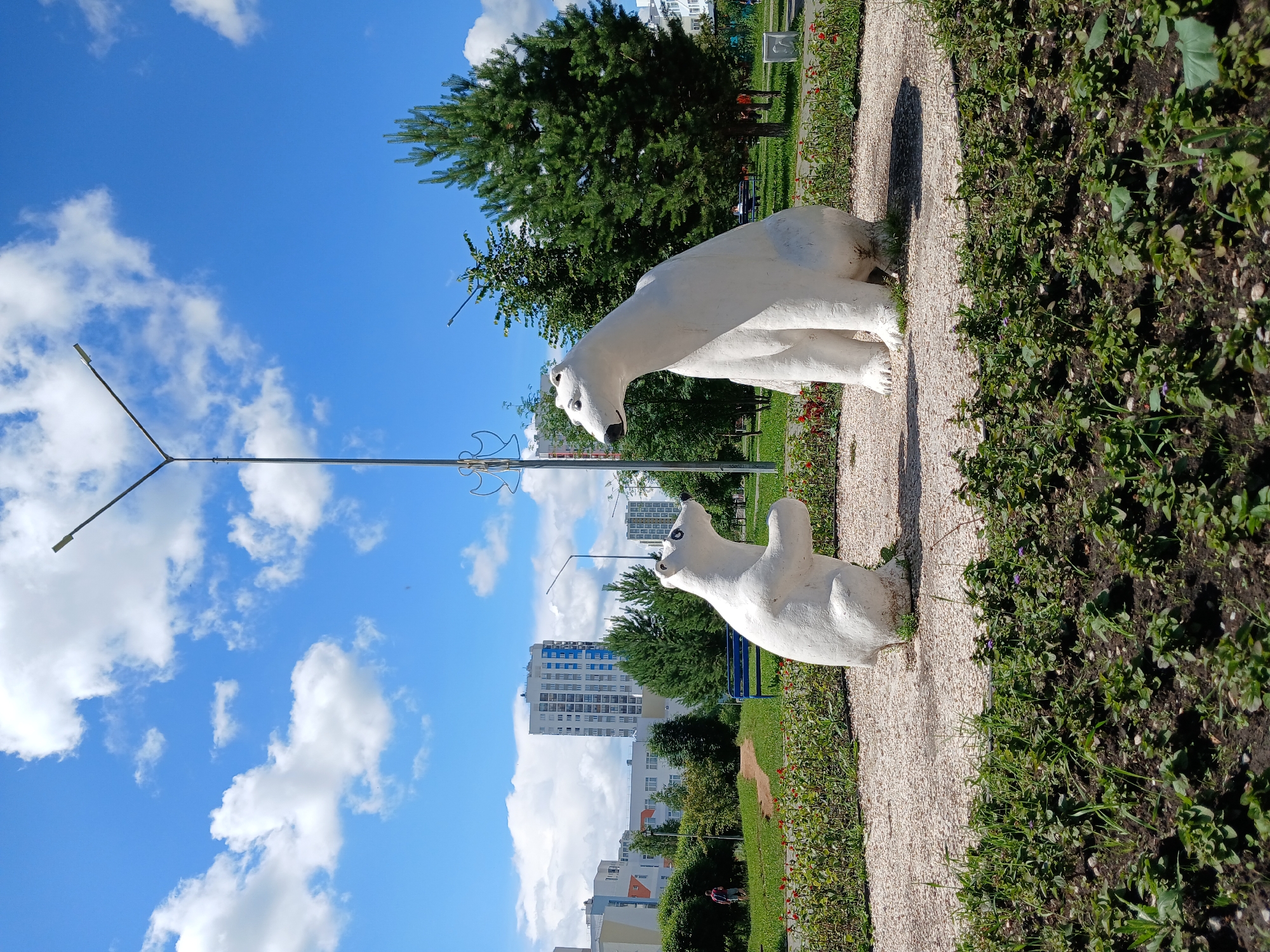 The image captures a sunny day in a park or public square in Yekaterinburg, Russia. Dominating the foreground are two white sculptures of polar bears. The larger bear is seated, facing to the right, while a smaller bear stands on its hind legs to the left, with its front paws raised as if interacting with the larger bear. Both sculptures are set on a bed of light-colored gravel. Around the gravel area, there are flower beds with green foliage and some red flowers.
In the midground, a path winds through a grassy area, and a blue park bench is visible. Beyond the park area, modern residential buildings with multiple stories and light-colored facades are present, some featuring blue accents. Dense green trees, including a prominent pine tree on the right, dot the landscape. A tall lamppost with multiple light fixtures stands centrally in the background. The sky is a vibrant blue with scattered white, fluffy clouds. A person wearing a backpack is visible walking on a path in the distance to the left, and a few other indistinct figures are present further back.