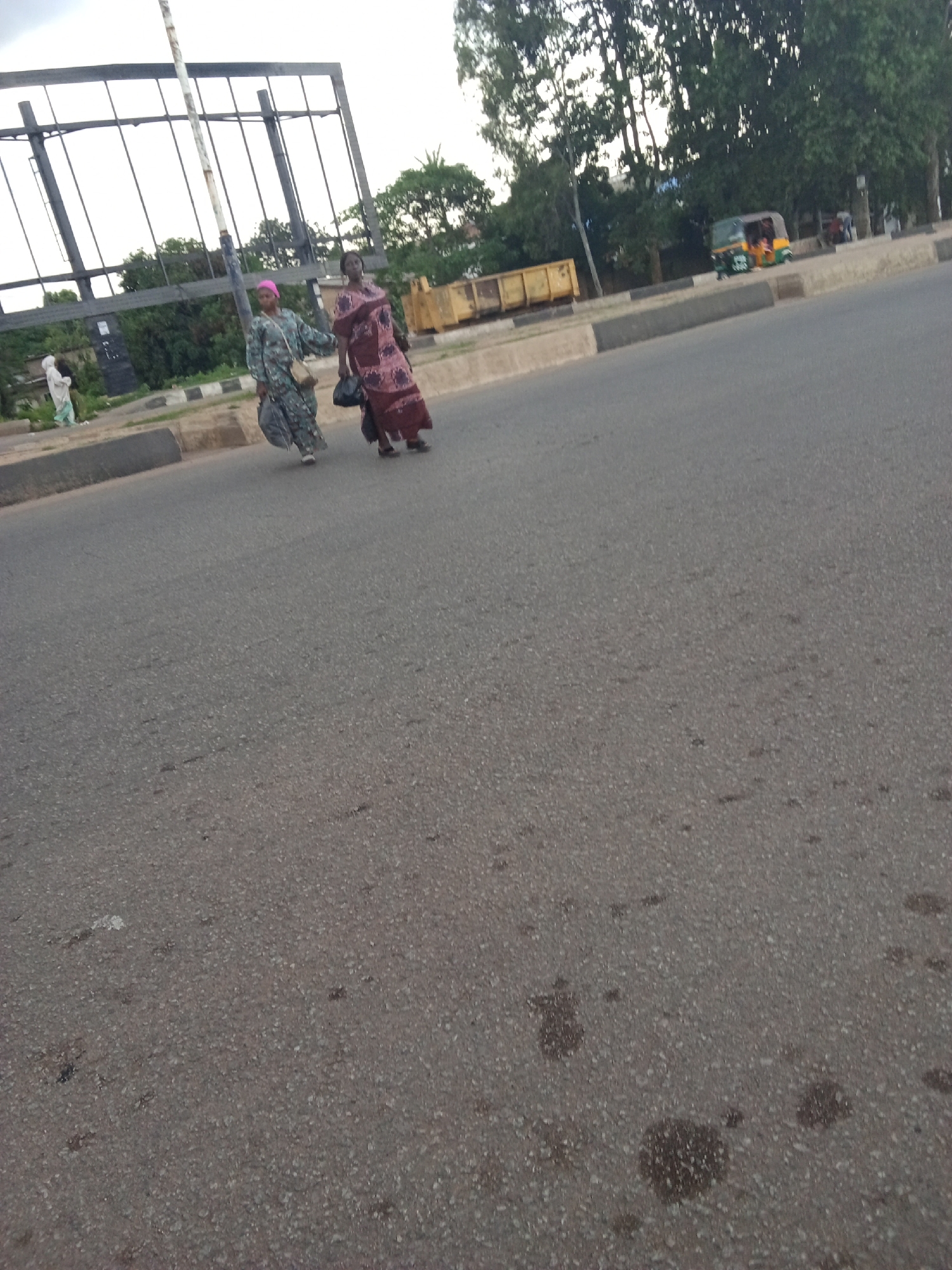The image depicts a street scene in Jos, Nigeria. Two women are walking on a paved road. The woman on the left wears a patterned blue and green dress and a pink headscarf, carrying a beige bag. The woman on the right is in a maroon and patterned dress, holding a black plastic bag. Another person in a white garment is visible further back on the left. In the background, there's a large metal structure, trees, a yellow construction container, and a small three-wheeled vehicle. The road appears to be a multi-lane thoroughfare with a concrete median barrier. The sky is overcast.