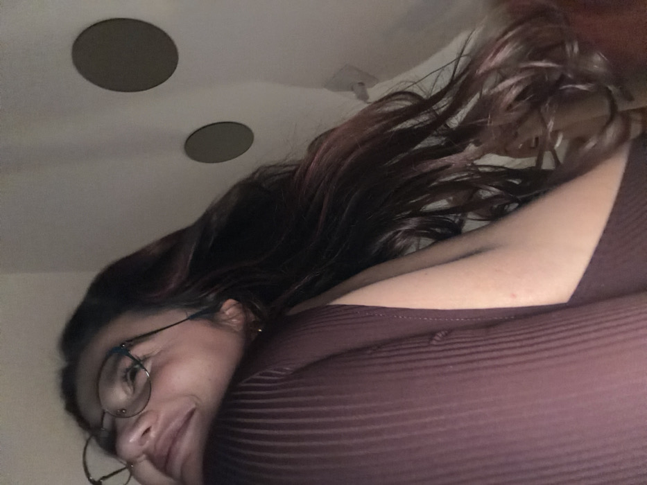 A woman with long, dark hair and glasses looks down at the camera. She's wearing a dark purple, ribbed top. The background is a white ceiling with two circular lights and a vent. The angle is from below, looking upwards at the subject. Her hair is partially obscuring her face and shoulder.