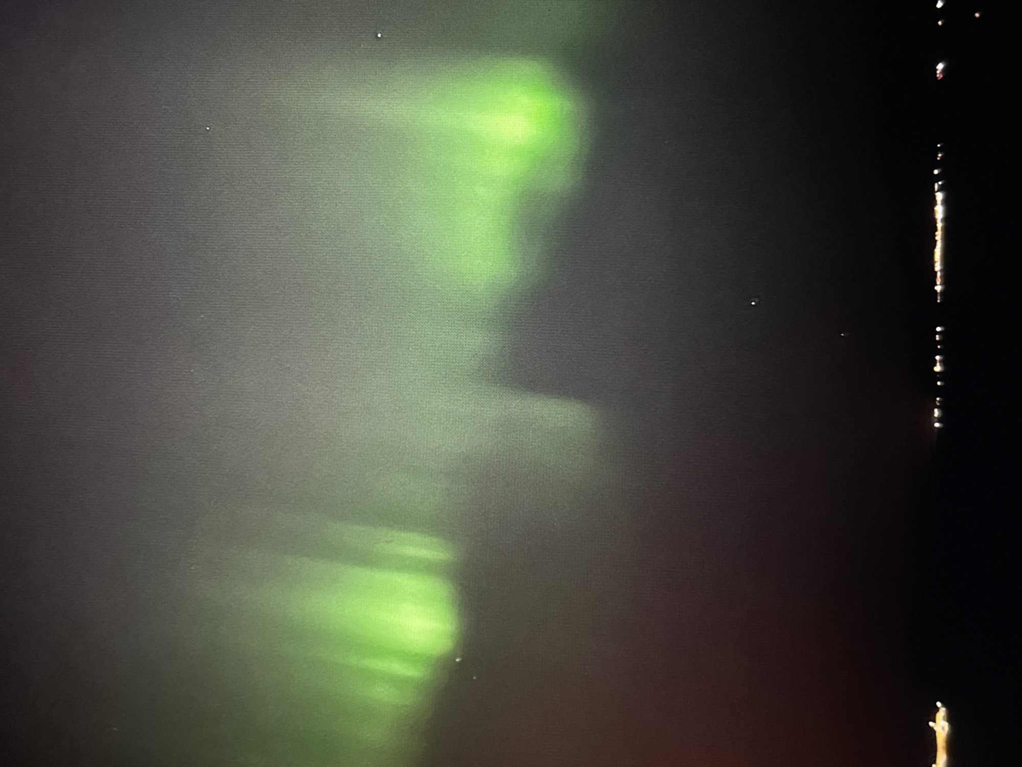 The image depicts a nighttime scene in Unknown, Unknown. A vibrant green light stretches vertically across the left side of the frame, appearing as an aurora or similar atmospheric phenomenon. The right side of the image is largely dark, with a vertical line of bright, scattered points of light, possibly reflections on water or distant lights. The overall environment seems to be dark with atmospheric interference or light phenomena.