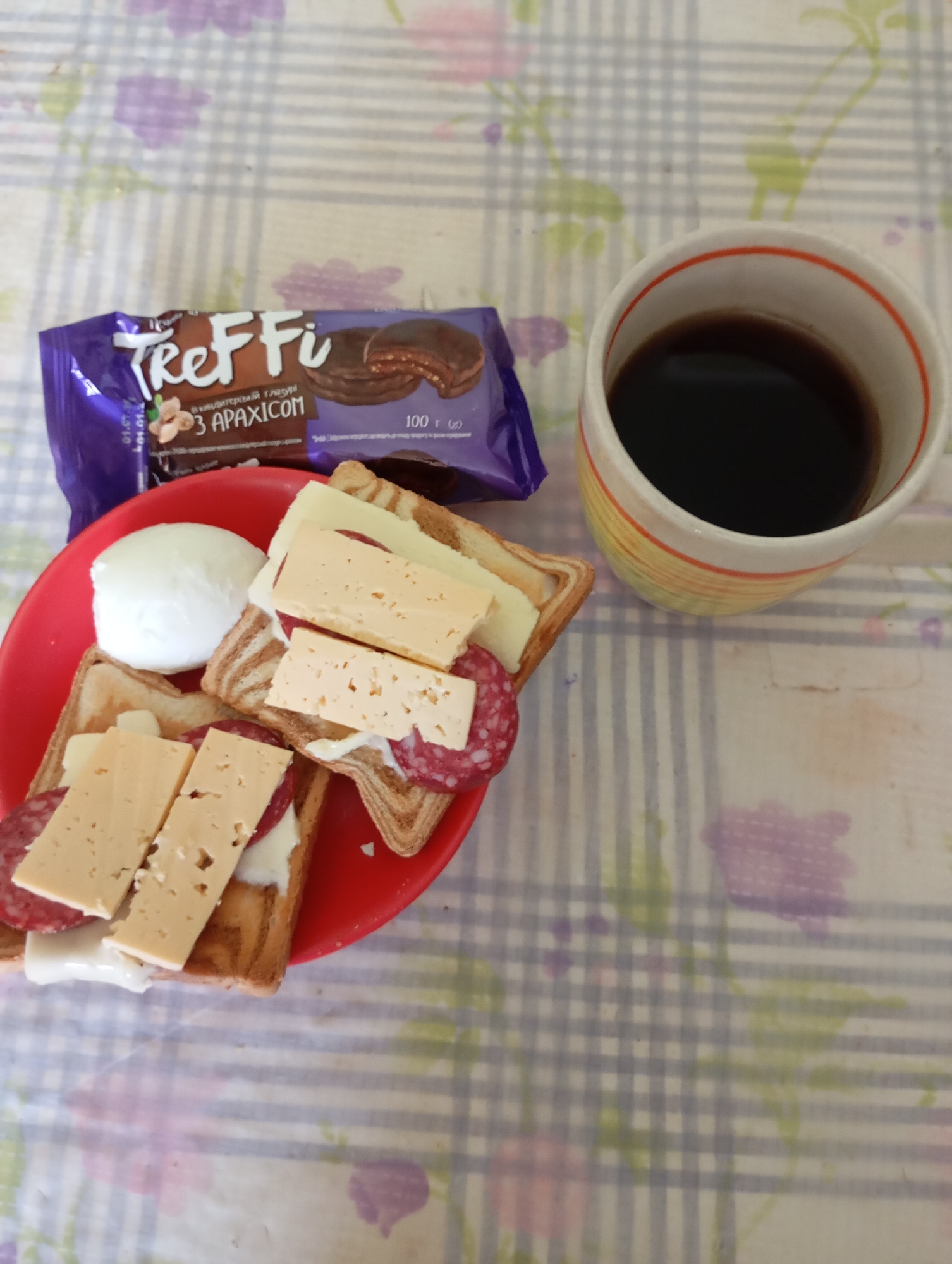 On a checkered tablecloth with a floral pattern, a red plate holds an open-faced sandwich made of toasted bread, sliced salami, and cheese. A hard-boiled egg is placed to the side of the sandwich. Next to the plate is a purple package of "Treffi" chocolate cookies. To the right of the plate, a white and yellow mug filled with dark coffee is positioned. The scene is set indoors, likely on a table, within a domestic environment in Horishni Plavni, Ukraine. There are no people present in the image.