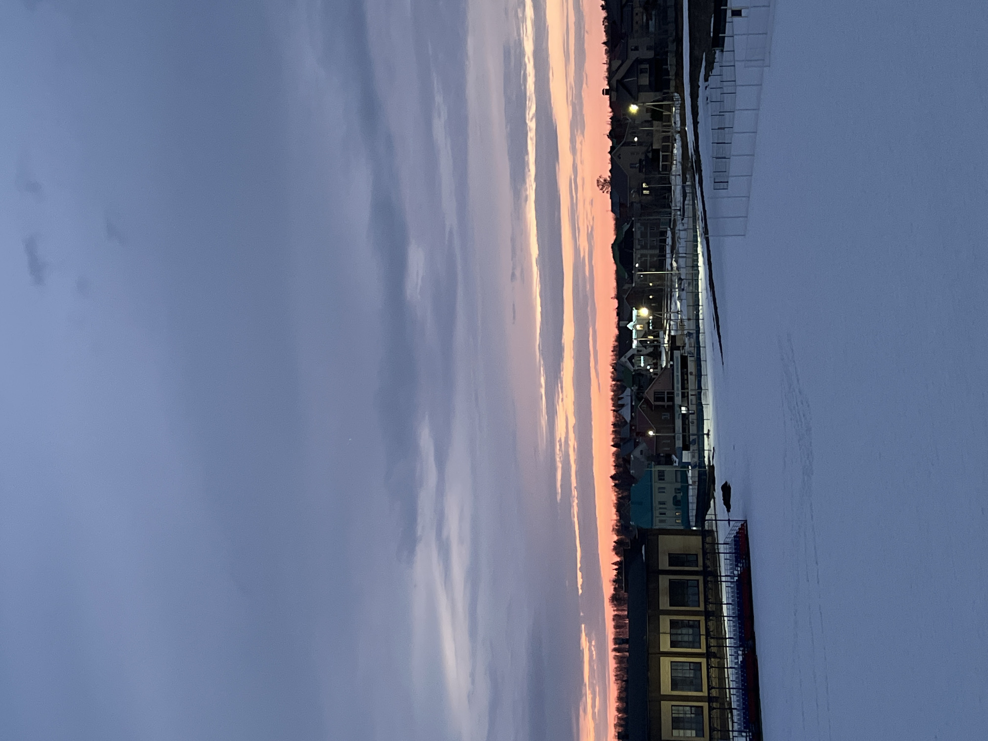 The image depicts a winter evening in Noviy, Russia. The foreground is covered in snow, suggesting a frozen river or lake. A stadium with seating is visible along the shoreline, with adjacent buildings. The sky transitions from a dark, cloudy blue to a pinkish-orange gradient near the horizon. Streetlights illuminate residential buildings in the background.