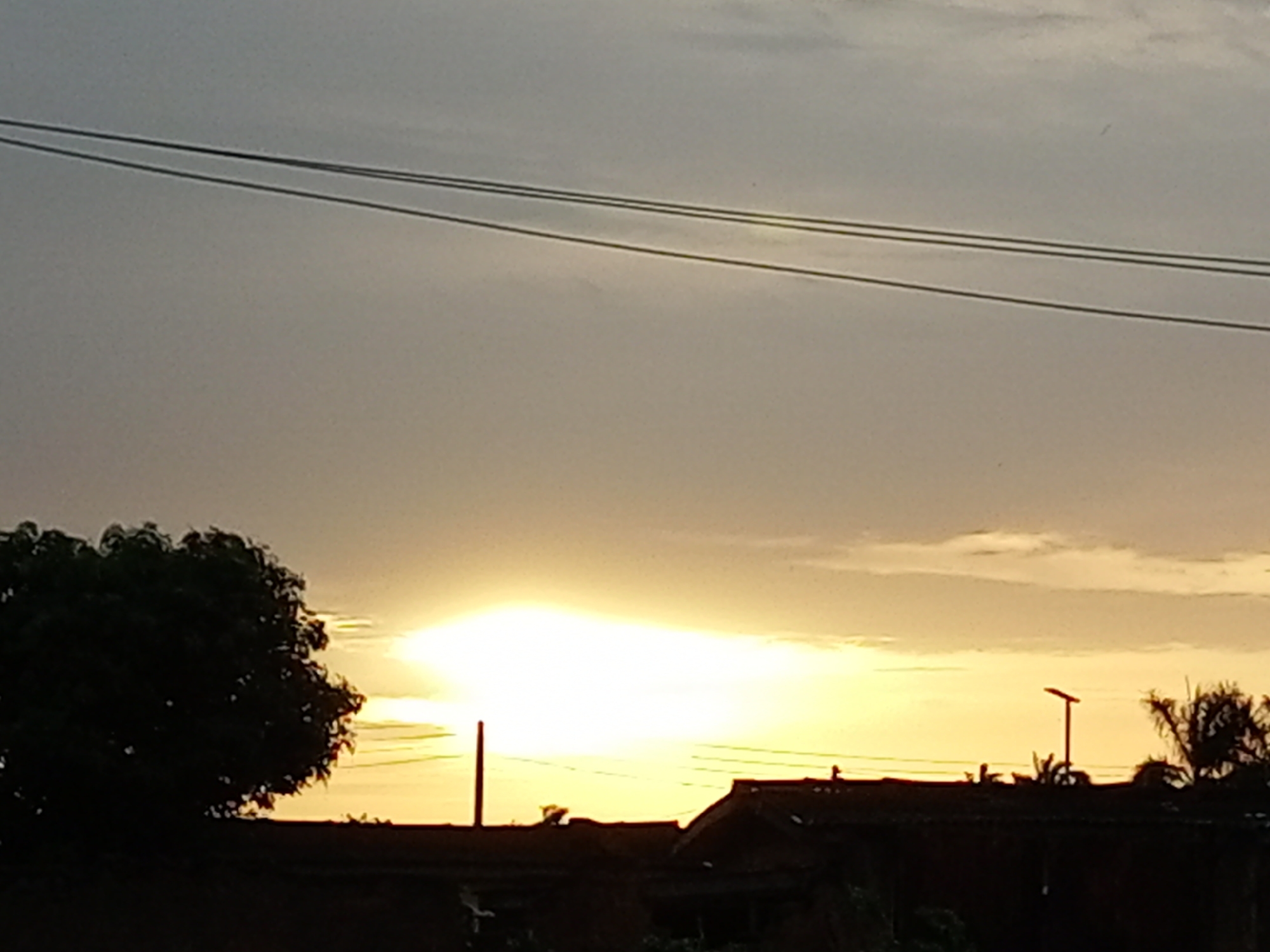 The image depicts a sunset scene in the city of Barden Yana, Nigeria. The sky is overcast with clouds, and the sun is a bright, diffused glow near the horizon. In the foreground, silhouetted against the sky, are the rooftops of several buildings. A large, dark tree is visible on the left side of the frame, and on the right, a silhouette of palm trees and a street lamp can be observed. Power lines are strung across the upper portion of the image. The overall impression is one of a calm, late afternoon or early evening atmosphere in a Nigerian urban setting.