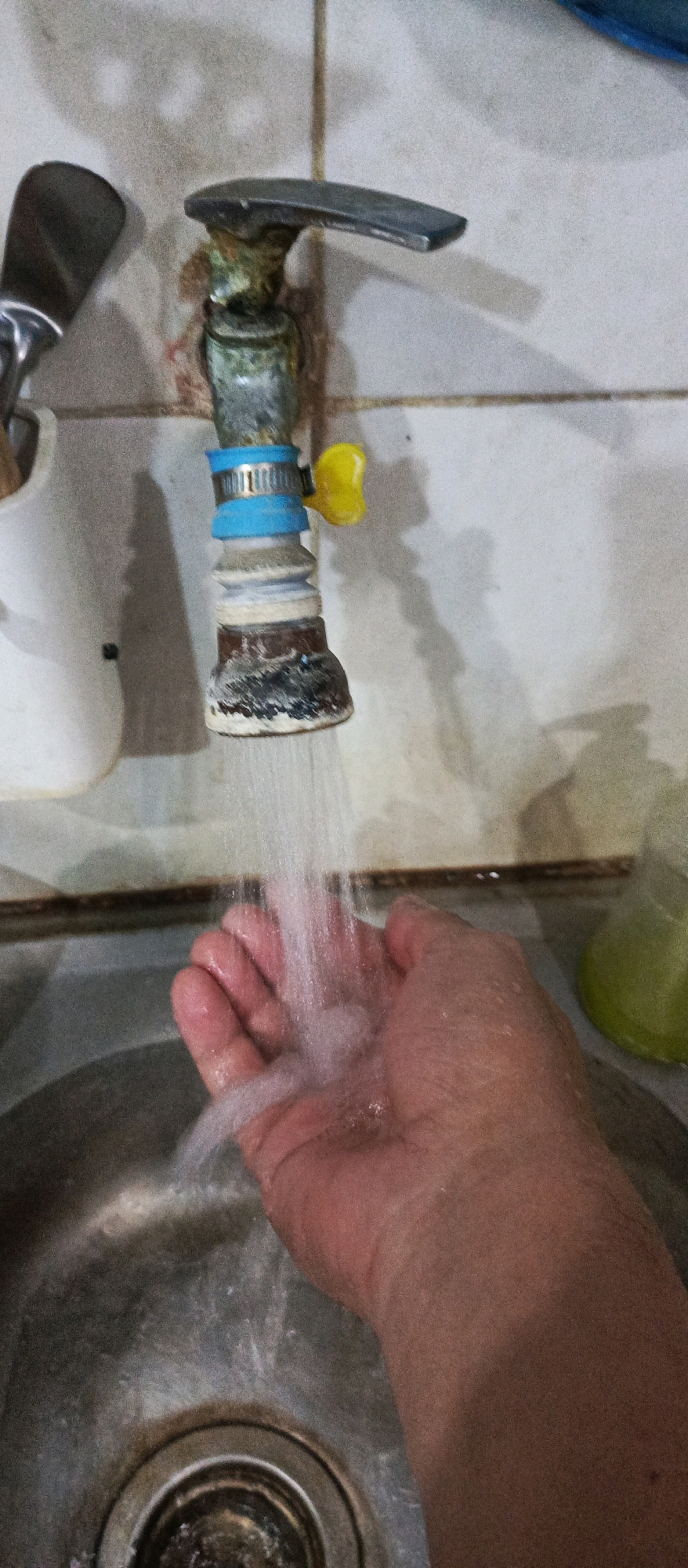 A close-up, eye-level shot shows a person's right hand under a dripping faucet in what appears to be a kitchen or bathroom sink area in Sidoarjo, Indonesia. The faucet, old and stained with mineral deposits, is made of metal with a silver handle and a yellow knob, a blue band with a metal clamp around the spout, and a brown lower section. Water streams from the spout onto the palm of the hand. The sink is made of stainless steel and shows signs of wear and grime. The background consists of a tiled wall, with a white container holding utensils to the left and a blurred green object, possibly a bottle, to the right. The lighting is dim, casting shadows on the wall.