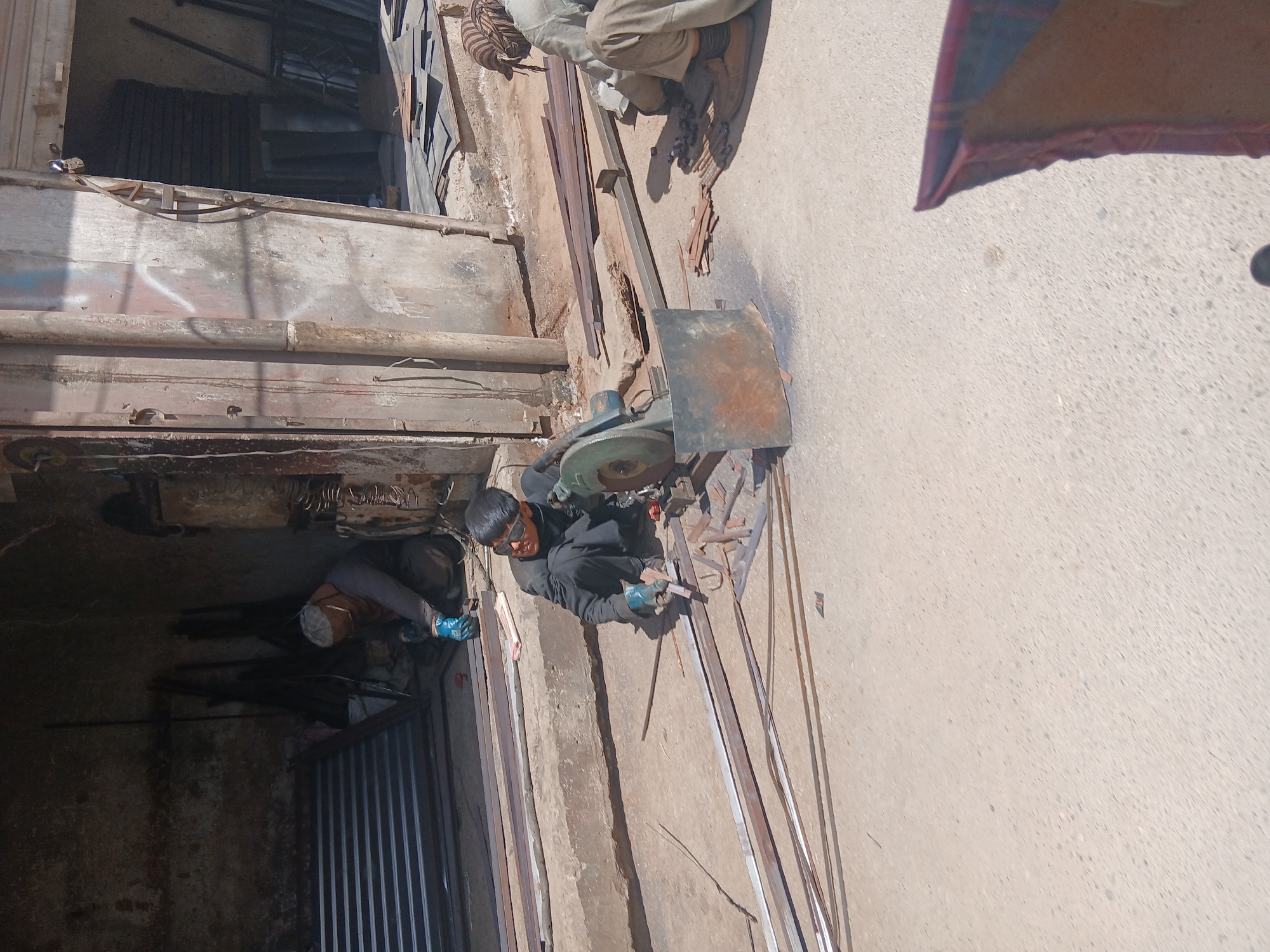 The image depicts a scene in Quetta City Tehsil, Pakistan. A man wearing safety goggles and gloves is using a chop saw to cut metal rods on a concrete surface outdoors. Another person, wearing a face mask and gloves, is partially visible in the background, inside what appears to be a workshop. The workshop has a corrugated metal door. Several metal rods are scattered on the ground around the man using the saw. A second person is seated near the wall adjacent to the first man cutting the metal. Debris and tools are around him.