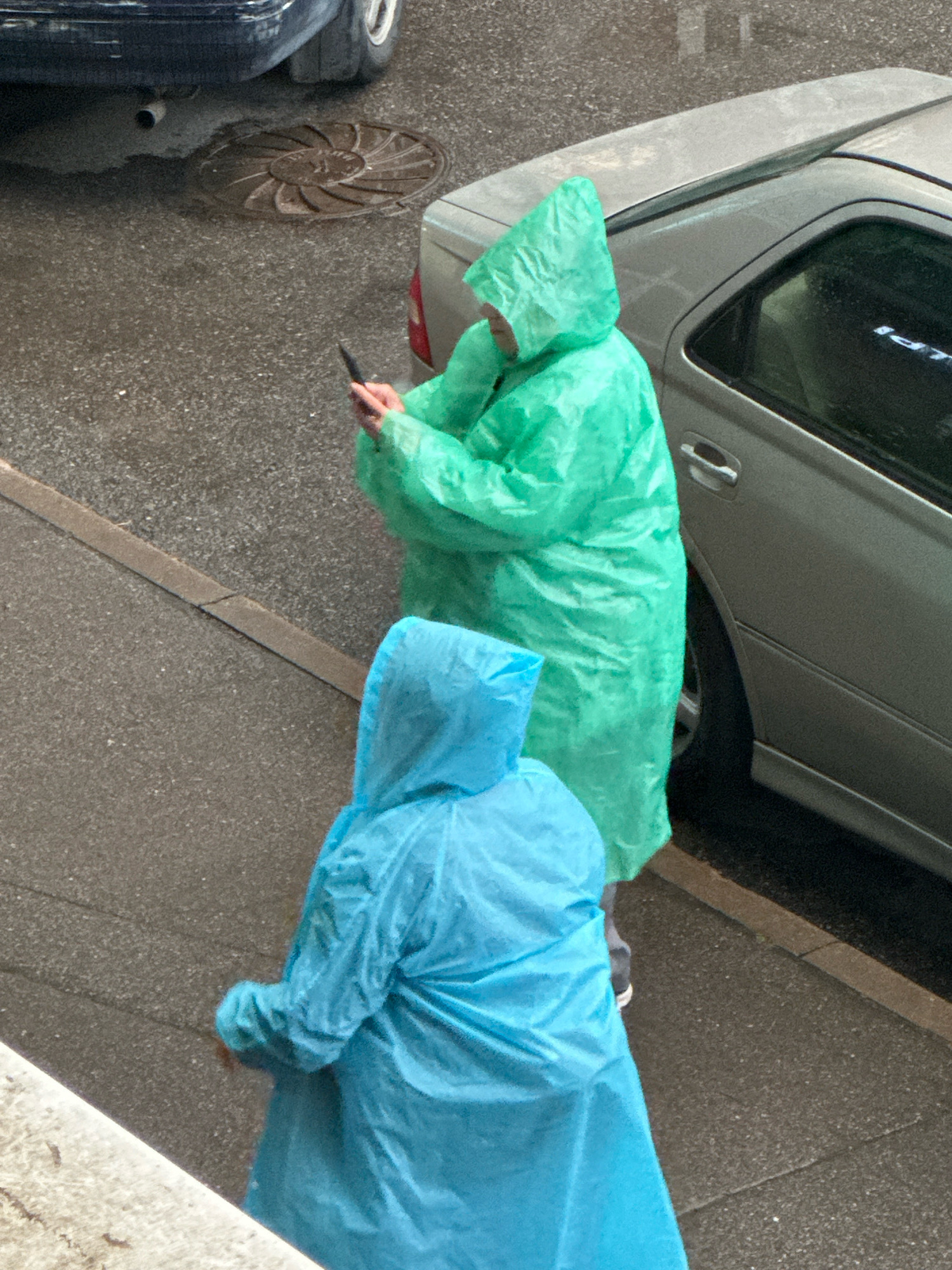 Two individuals wearing hooded raincoats, one bright green and the other bright blue, stand on a wet asphalt surface. The person in the green raincoat is positioned behind and to the right of the person in the blue raincoat, and is holding a black smartphone in their right hand. They are both facing away from the camera. A silver car is parked to the right of the individuals, partially obscuring the view of the green-raincoated person. To the left of the individuals, a portion of a dark-colored vehicle is visible, along with a manhole cover in the asphalt. The overall scene suggests an outdoor environment, possibly a street or sidewalk, in a city. The presence of raincoats and wet ground indicates inclement weather. The lighting is somewhat dim, typical of overcast conditions.