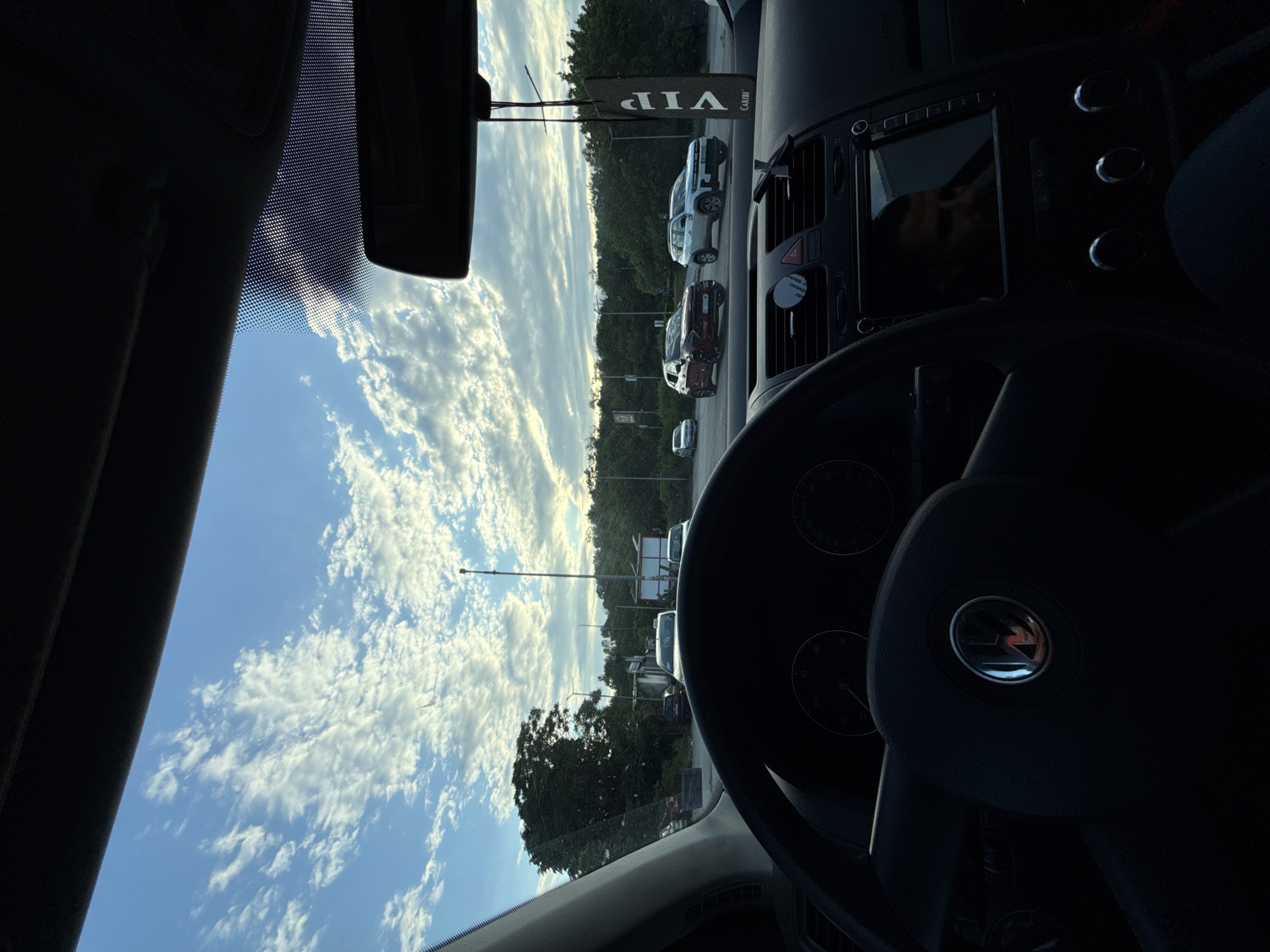 Captured from inside a car, the scene shows a sunny sky with scattered clouds visible through the windshield, complemented by street views of Lovech, Bulgaria, based on the text "VIP" in a blurry sign hanging from the rearview mirror. Several vehicles are parked along the roadside. The steering wheel of the car is prominently displayed in the foreground, obscuring a clear view of the dashboard and infotainment system. The interior is dimly lit, contrasting with the brightly illuminated exterior.