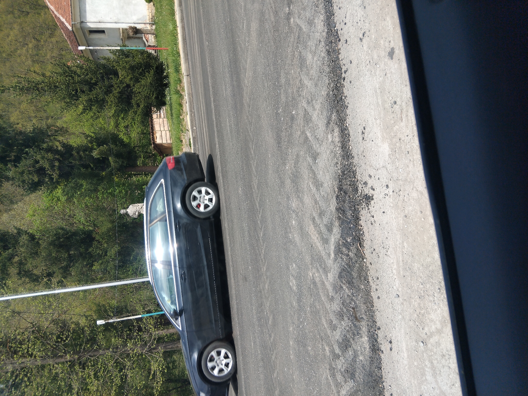 The image captures an outdoor scene in bright daylight, rotated 90 degrees clockwise from its natural orientation. A dark-colored sedan, possibly an Audi, is parked on the right side of a paved asphalt road. Its right wheels are on a gravel and dirt shoulder, while the left wheels are on the asphalt. Sunlight brightly reflects off the car's windows.
To the left of the road, a verdant landscape of numerous trees and bushes dominates the background, indicating a rural or semi-rural setting. In the upper left portion of the image, behind the foliage, a house with a reddish-brown tiled roof and light-colored walls is partially visible.
A notable feature is a tall white pole standing near the house, adorned with red, white, and green stripes at its base. The word "VELCHEVO" is clearly inscribed on this pole. Further amidst the trees, a light-colored, human-like figure, possibly a statue or scarecrow, is faintly discernible. Another slender white pole, potentially a utility pole, is visible in the middle-left ground, with overhead lines stretching across the upper frame.
The weather appears clear and sunny, as evidenced by the strong reflections and lack of clouds. No people are visible, and the scene depicts a static, quiet moment. The visible text "VELCHEVO" directly points to the location context of Velchevo, Bulgaria.
