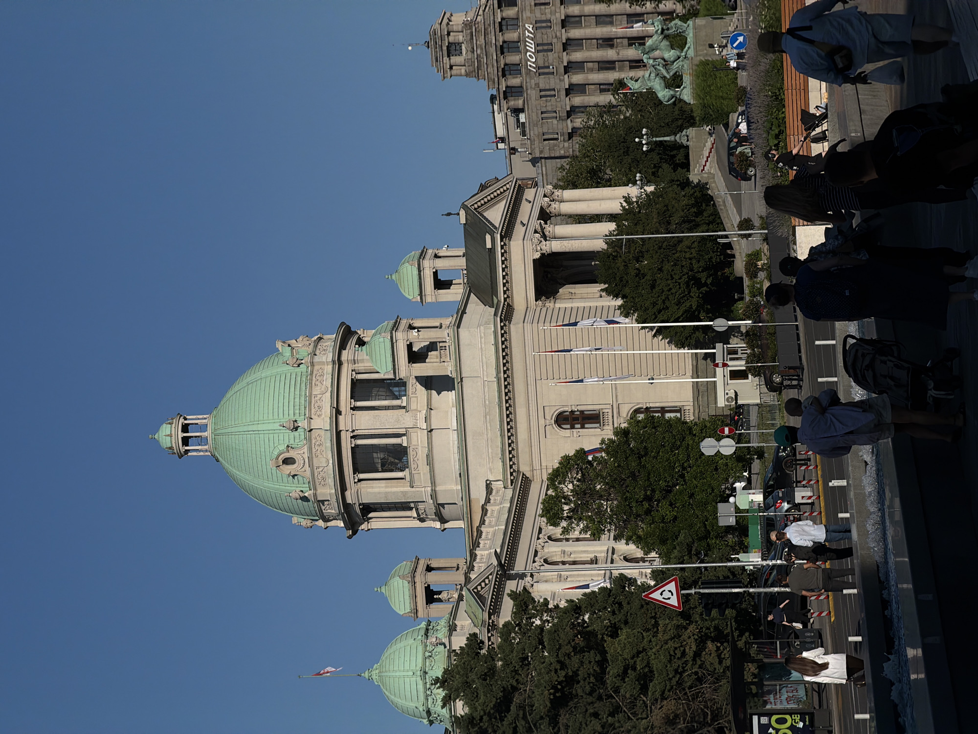 The image depicts a sunny daytime scene in front of the National Assembly of Serbia building in Belgrade, Serbia. The grand building features a large central dome and multiple smaller domes, all painted a distinctive green. Classical architectural elements, including columns and ornate stonework, are prominent. Several flags, including the Serbian flag, are visible on poles.
In the foreground, a group of people are walking and standing near a low wall, with a body of water or fountain in front of them. Some are pushing strollers. Beyond the wall is a street with cars and more people. On the right side of the image, a statue of a horse and rider is visible in front of another imposing stone building, which has "ПОШТА" (meaning "Post Office" in Cyrillic) inscribed on its facade. Trees and streetlights are also present, contributing to the urban environment. The sky is clear blue.