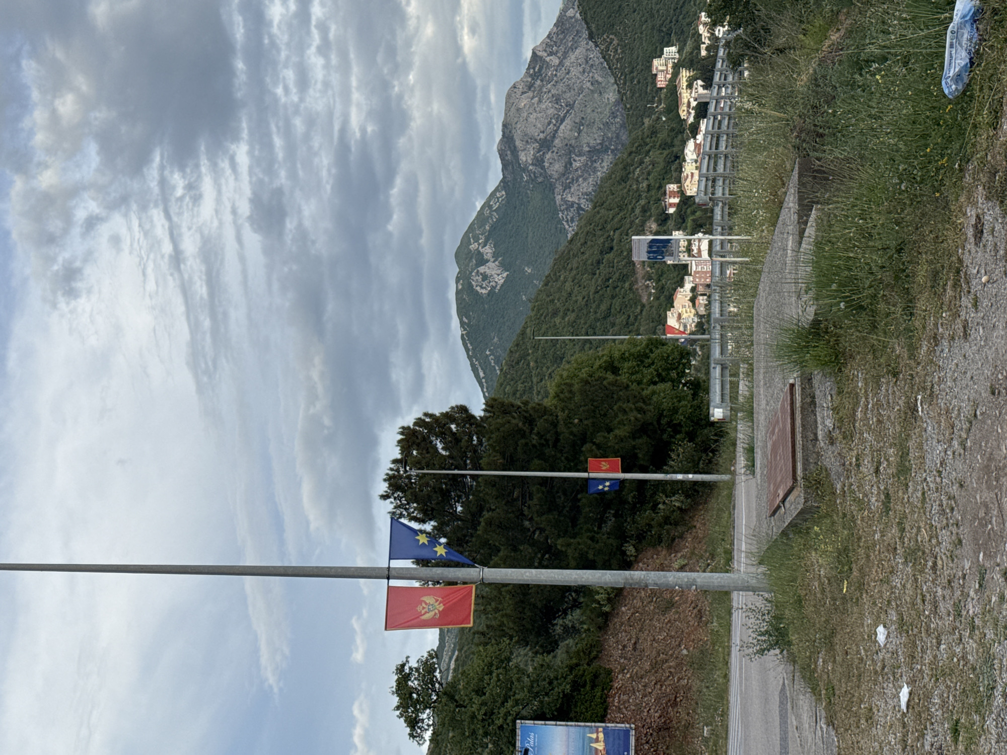 The image shows a roadside scene near Medinski Krš, Montenegro. The foreground features a metal pole displaying the flags of Montenegro and the European Union. Beyond the pole are trees and vegetation. Further in the background is a mountain, with buildings visible on its slopes. The sky is overcast with layered clouds. A paved road runs alongside the grassy shoulder in the lower right corner of the frame.