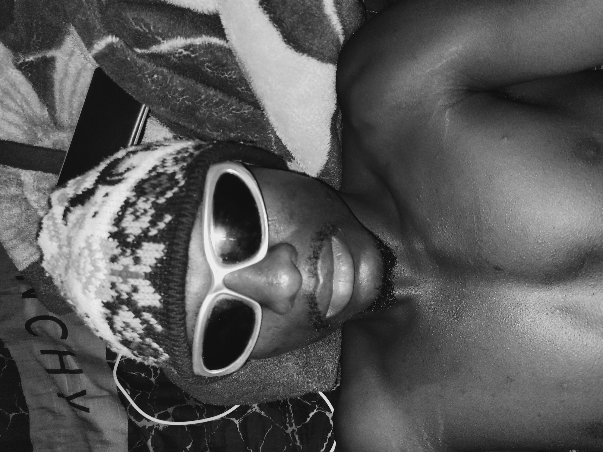 A black and white, tilted, close-up shot shows a man lying down with a knitted hat and sunglasses, partially shirtless, on what appears to be a patterned fabric or bed. A dark rectangular object, possibly a phone or small tablet, is to the left of the man's head. A white cord is visible near his head. The location is not discernible from the image and cannot be specified as Bukuru, Nigeria.