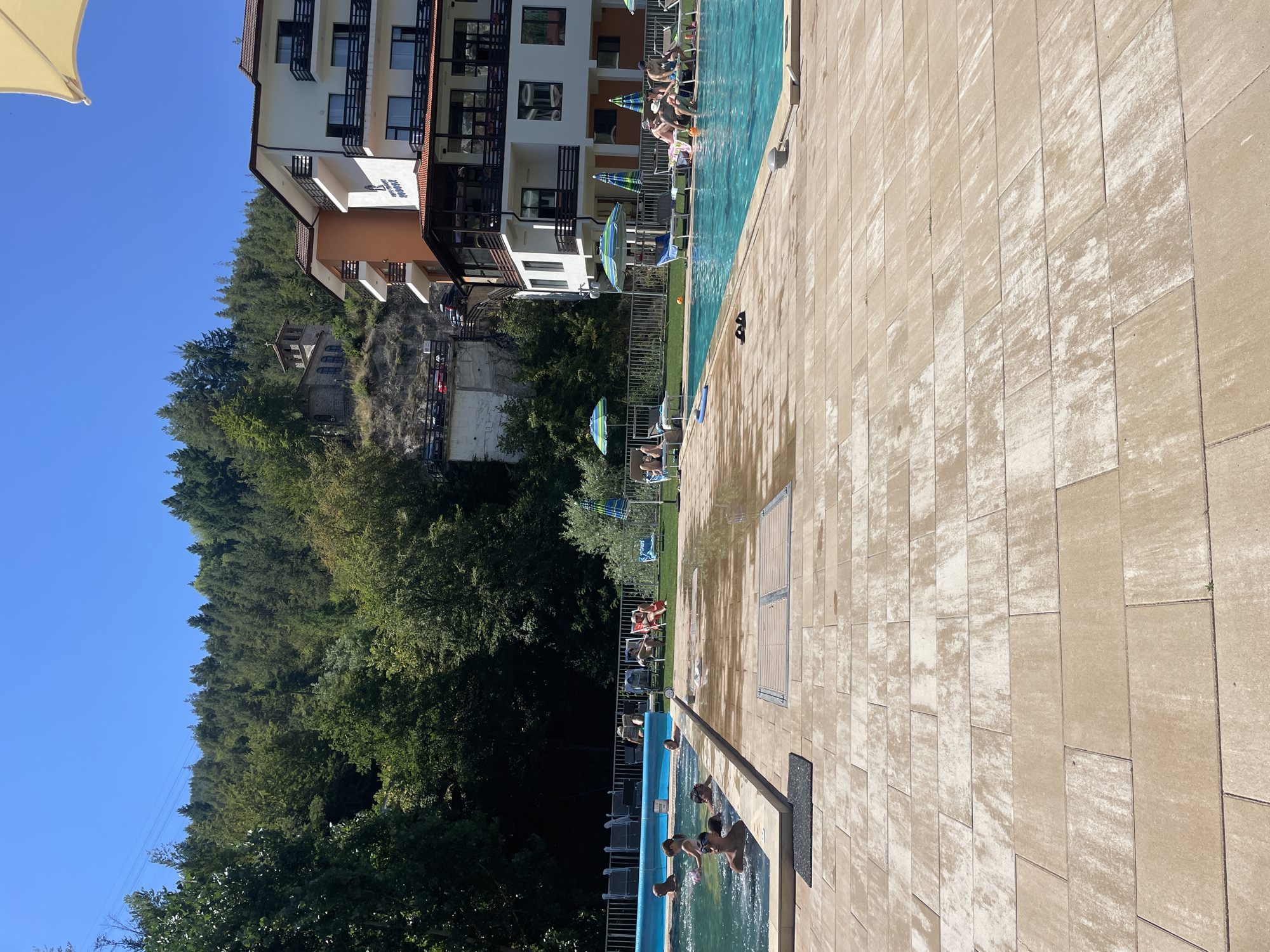 The image depicts a sunny day at an outdoor swimming pool complex in Banite, Bulgaria. The foreground is dominated by a tiled patio area leading to a clear blue swimming pool. Several people are visible in the pool, some of whom appear to be children, engaging in recreational activities. On the poolside, lounge chairs are arranged, with a few individuals relaxing. In the background, a multi-story hotel building with balconies and a white facade stands prominently, adjacent to a hill covered in dense green forest. A historic stone building with a steeple is perched higher up on the hill. The sky is a clear, bright blue.