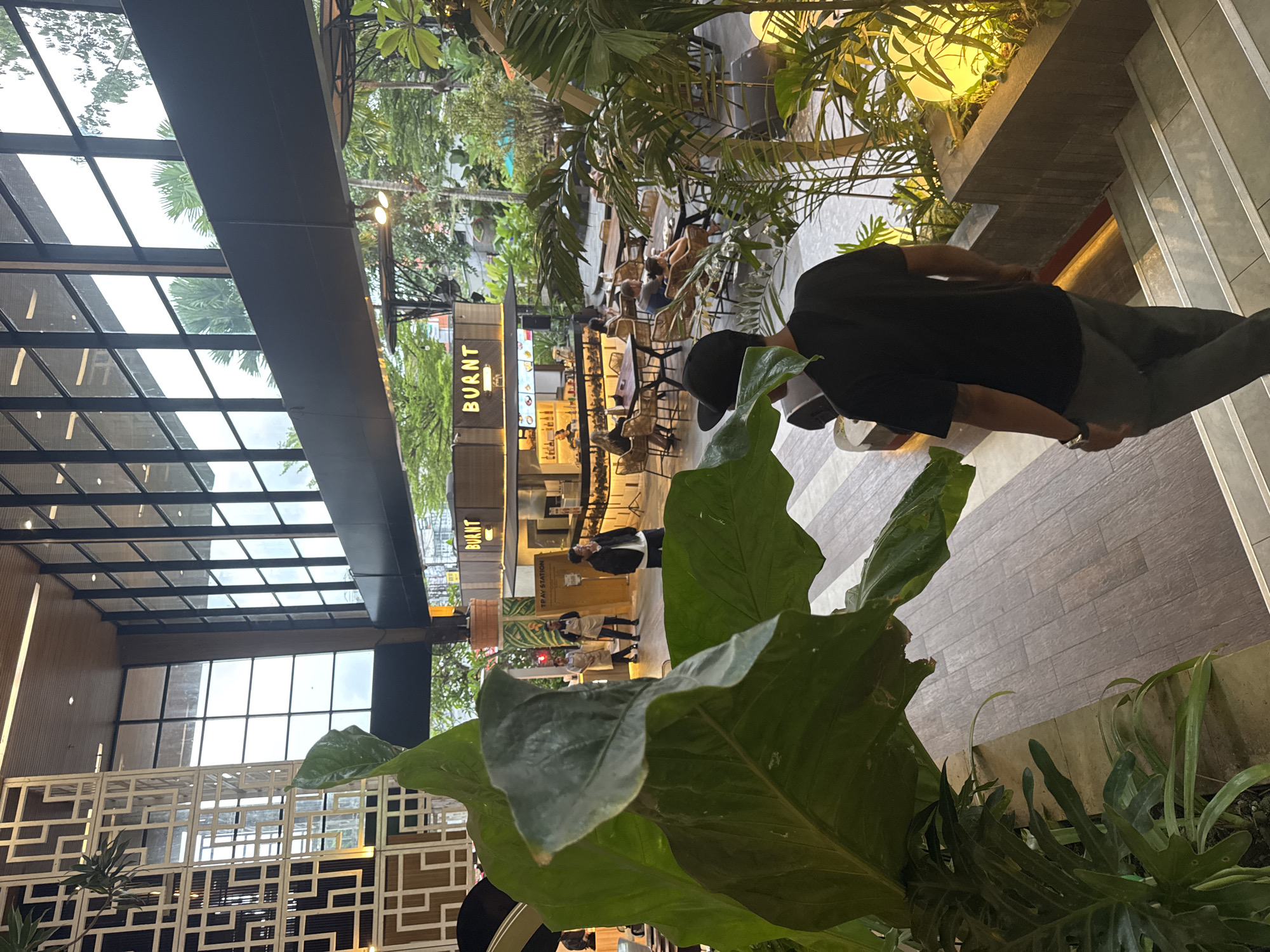 An indoor-outdoor commercial space, likely a food court or market, is captured in a portrait orientation. In the immediate foreground, a large, dark green, glossy-leafed plant partially obscures the view. To the right, a person in a black t-shirt, dark pants, and a black baseball cap is seen from behind, walking away from the camera down a set of stairs or a sloped walkway. This individual wears a watch on their left wrist.
The setting features modern architecture with a high, angular glass roof structure allowing natural light to filter in. Lush tropical foliage, including palms and other large-leafed plants, is integrated throughout the space in planters, creating a vibrant, green atmosphere.
In the mid-ground and background, several food stalls or small shops are visible. One prominent stall has the word "BURNT" displayed in white letters against a dark background. Another sign, partially obscured by plants, reads "TERAK STATION." Tables and chairs, often made of woven material, are arranged for dining, and several people are seated, some appearing to be eating or socializing. Other individuals, possibly staff, are visible near the stalls.
The flooring consists of textured, light-colored tiles. The overall impression is a casual, relaxed daytime environment, consistent with a tropical location.