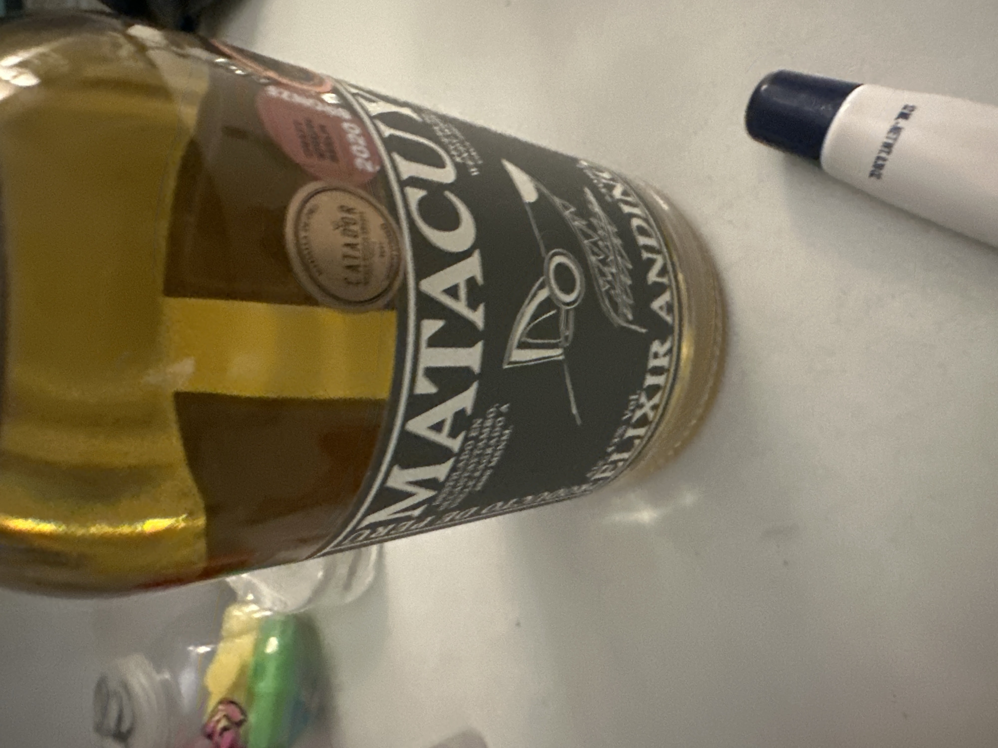 The image captures a close-up, angled view of a bottle of spirit and other objects on a light-colored surface, likely a table or countertop. The main subject is a clear glass bottle filled with a golden-yellow liquid, positioned horizontally. Its dark grey label features prominent white text, including "MATACUY" in large letters, with "LICOR DE HIERBAS ANDINAS" (Andean Herb Liqueur) and "PRODUCTO DE PERU" (Product of Peru) in smaller print below. Vertically aligned on the right side of the label is "ELIXIR ANDINO" (Andean Elixir). Two circular stickers are visible on the left side of the label; one is a "CATADOR" (Taster) award, and the other shows the year "2020."
To the upper right of the bottle is a white tube with a dark blue cap, bearing partially visible text that appears to read "DR. NEWT..." The foreground at the bottom of the image shows blurry, colorful objects, possibly candies or small items, enclosed within a clear plastic container or bag. The background is indistinct and brightly lit, suggesting an indoor setting. No people or specific activity are visible. The time of day is not discernible. The product's origin, Peru, is clearly indicated on the bottle label.